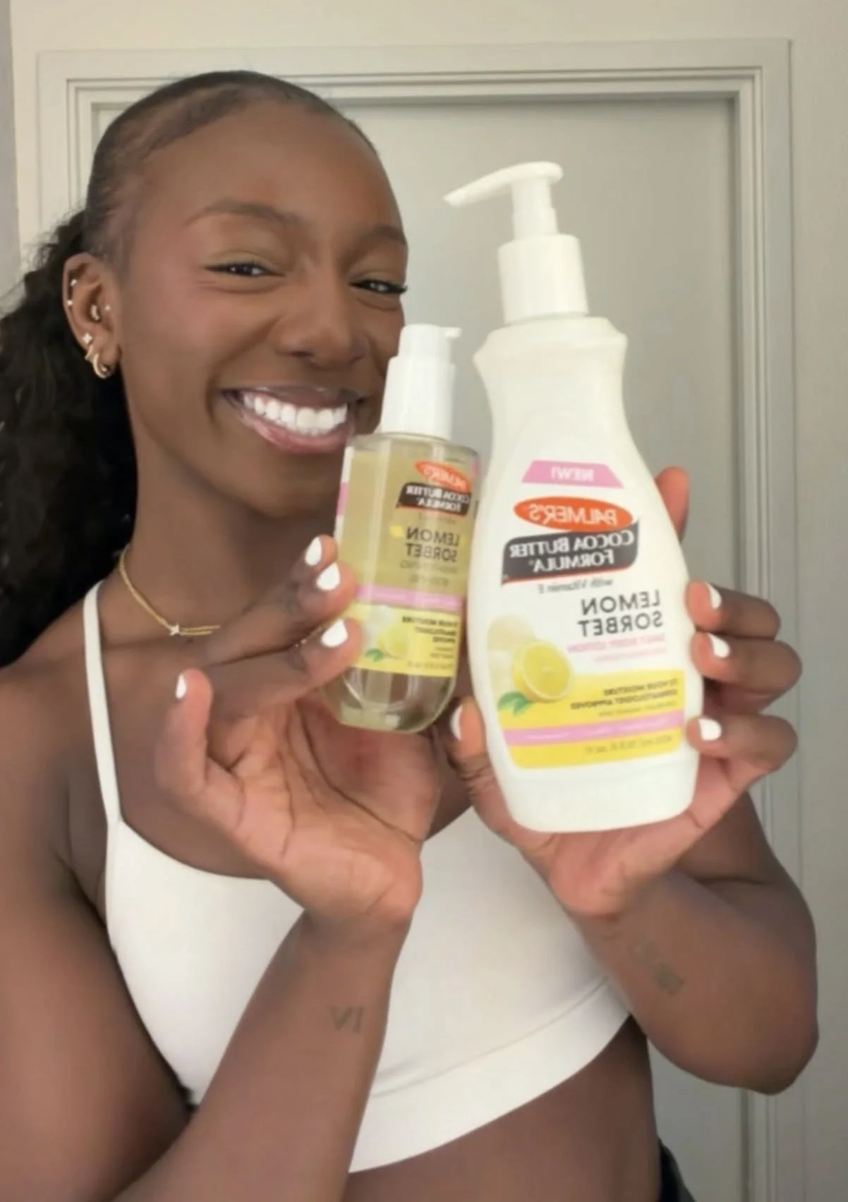 A woman smiling and holding various skincare products, including a bottle of lemon-scented foam soak and a bottle of lemon-scented foam scrub, in front of a door.