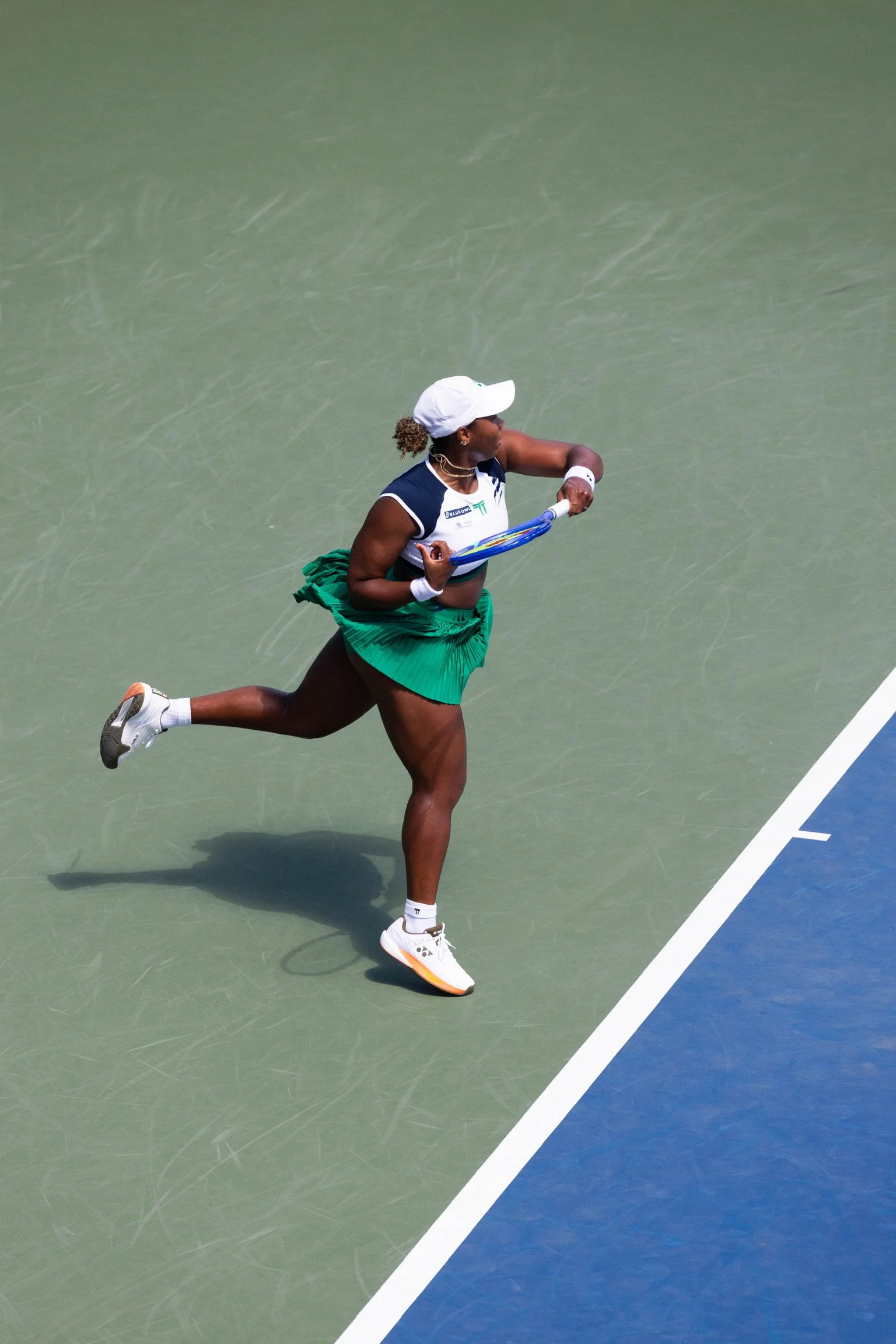 A female tennis player running on a green tennis court with a blue outer court boundary. She is wearing a white cap, a navy and white sports top, a green skirt, and white tennis shoes. She is holding a tennis racket in her right hand.