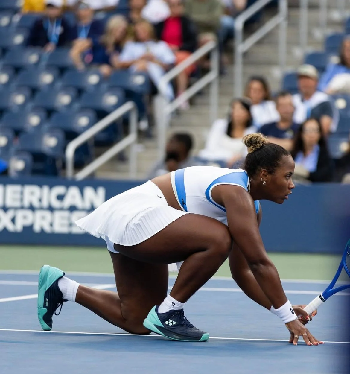 A female tennis player in a crouched position on the court, wearing sportswear and holding a racket, with spectators watching in the background.
