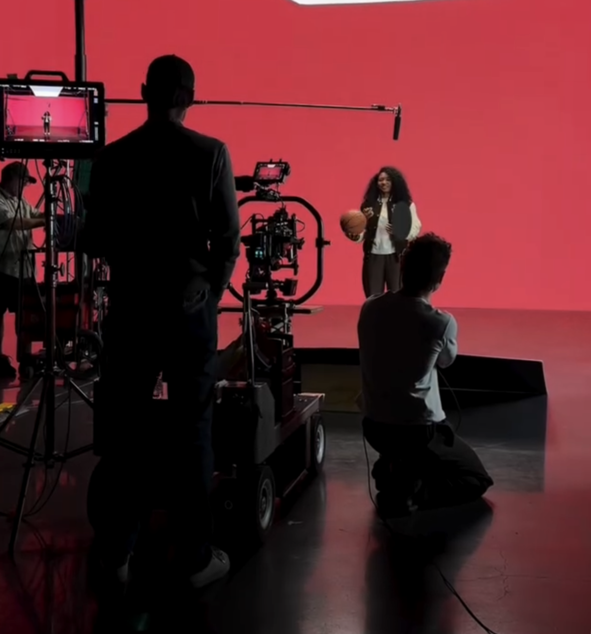 Behind the scenes of a video shoot with a woman holding a basketball and a phone, standing against a pink background, while crew members operate cameras and equipment.