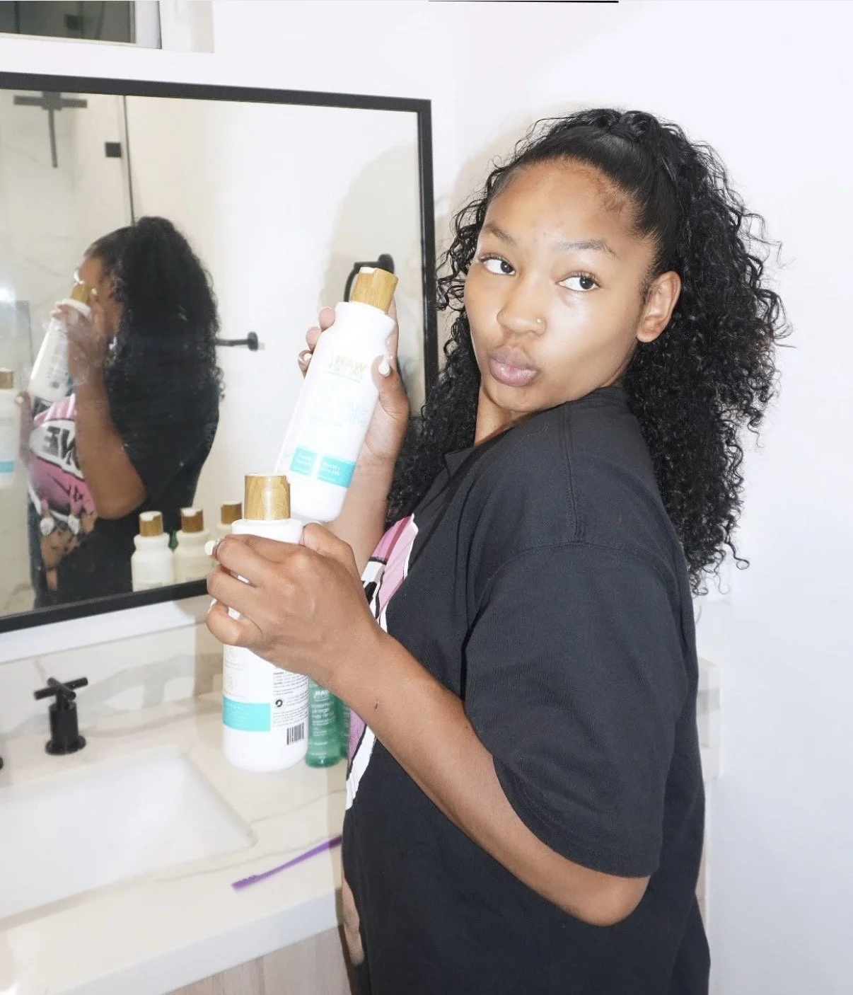 Young woman with curly hair holding skincare bottles in front of mirror in bathroom.