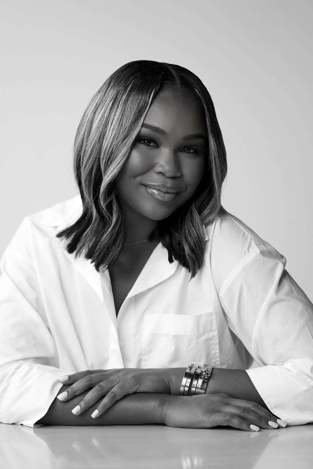 Black and white portrait of a woman with shoulder-length wavy hair, smiling, wearing a white blouse and bracelets, sitting at a table with her arms crossed.