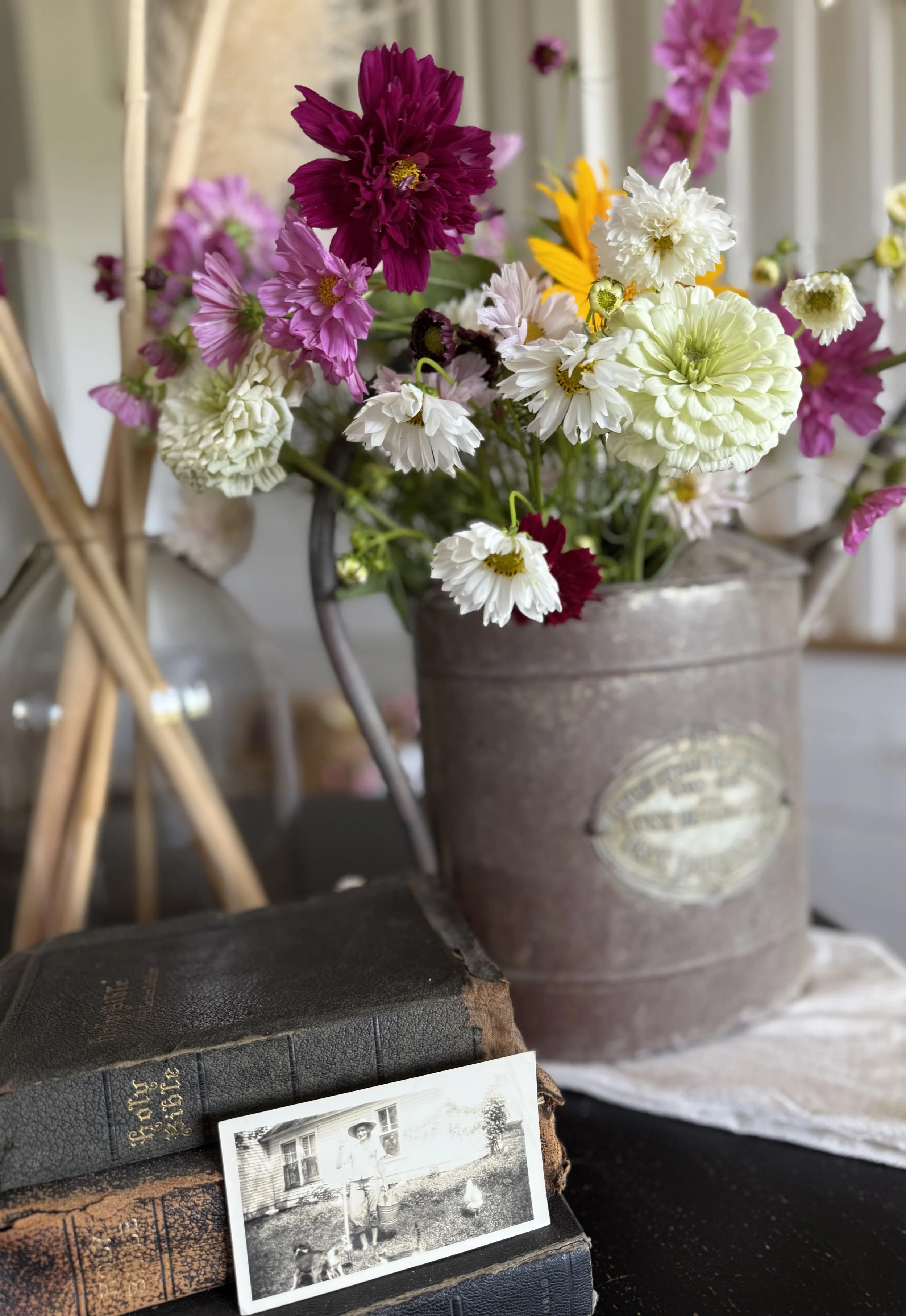 A vintage metal container filled with a colorful bouquet of flowers, including white, pink, purple, and yellow blossoms. In front of the container, there are old, worn books stacked with a black-and-white photograph resting on top, showing a person in vintage clothing standing outside a house with a dog nearby.