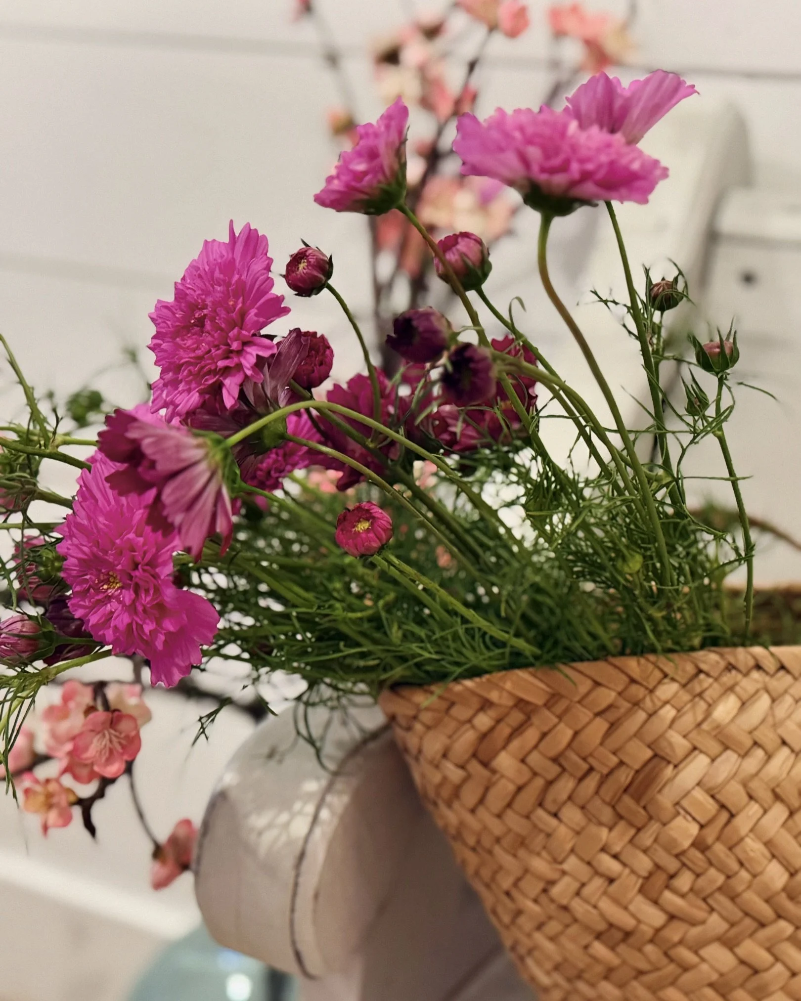 Pink and purple cosmos flowers in a woven basket on a white table with a blurred background of pink flowers.
