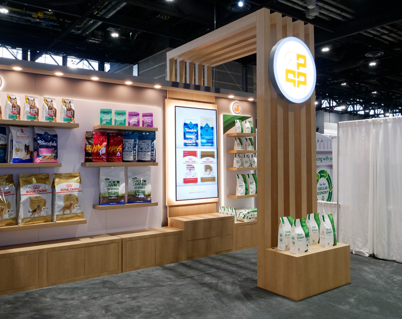 Trade show booth displaying various pet food bags on shelves, digital screens, and a round illuminated sign with a stylized logo on a wooden frame.