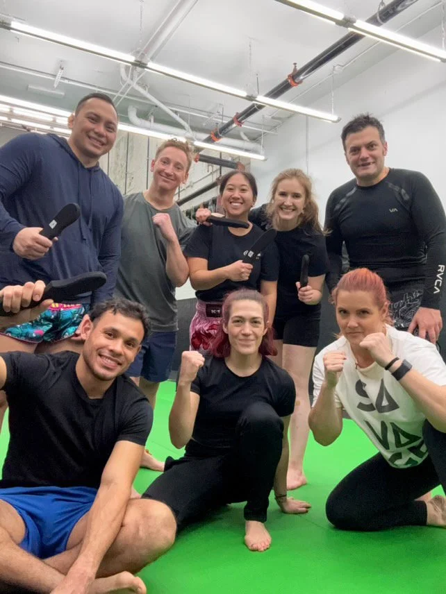 Group of eight people in athletic clothing posing for a photo in a fitness studio, some holding workout utensils, smiling and showing fist gestures, with a green mat on the floor.