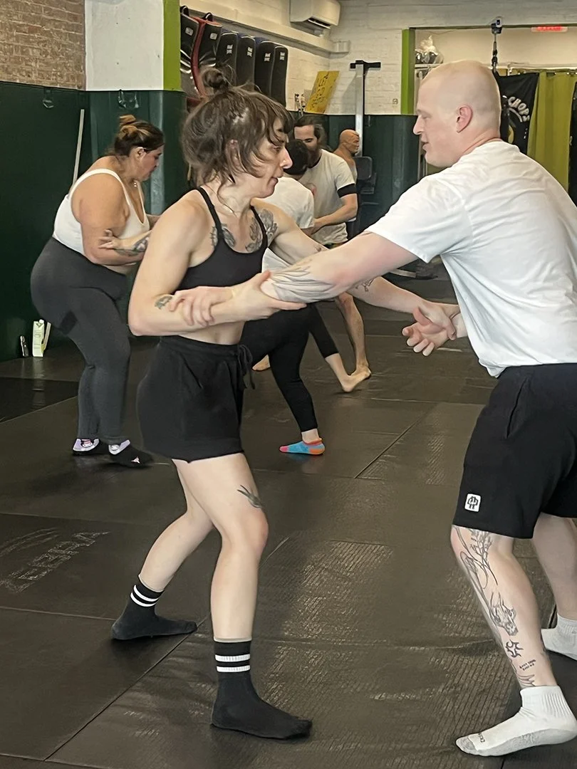 People practicing self-defense or martial arts in a gym, with a woman in black clothing being assisted by a man, others in the background engaging in similar activities.
