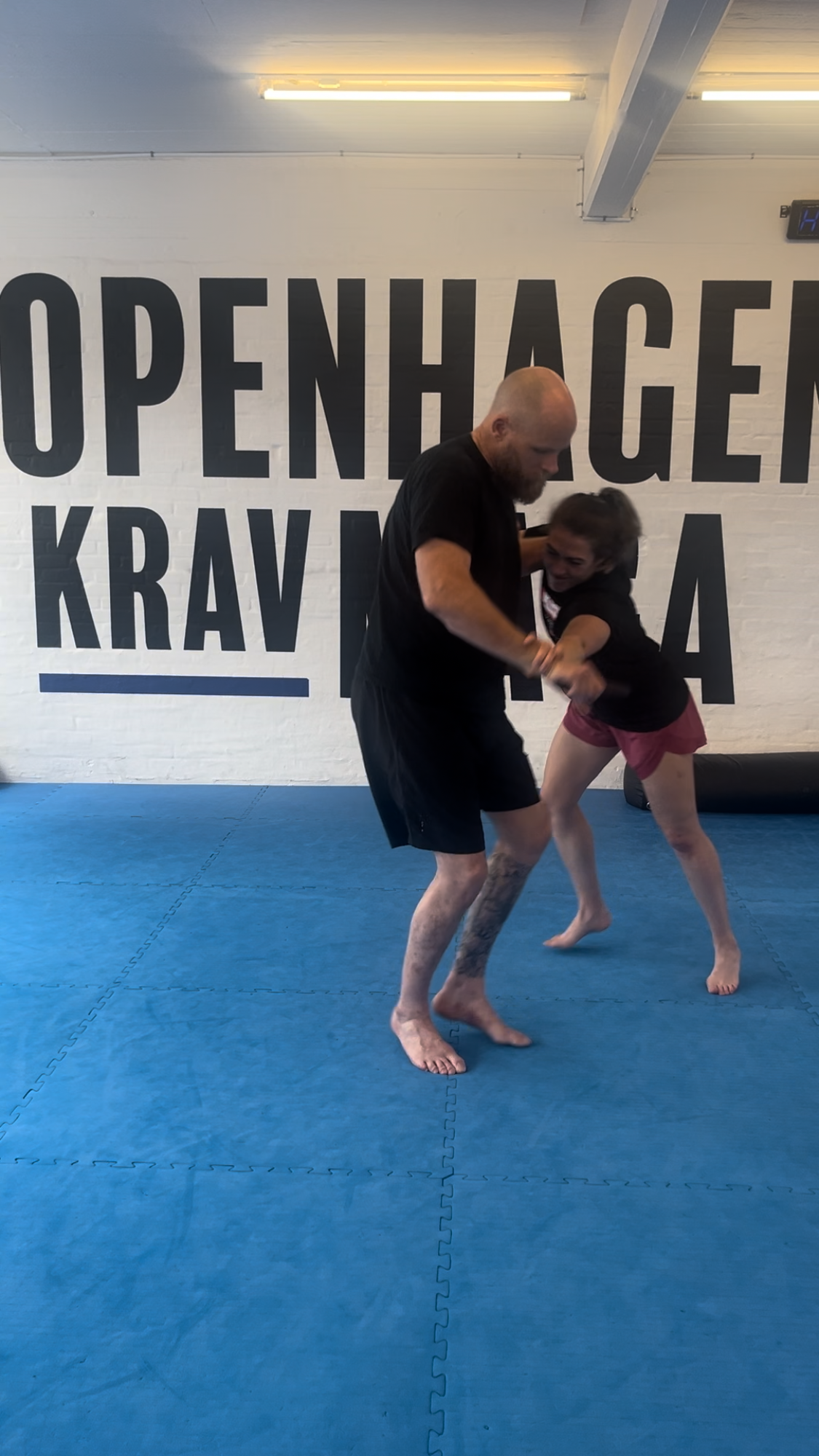 Two people practicing Brazilian Jiu-Jitsu on blue mats in a training facility with a large wall sign that says "OPEN H GENE KRA V".