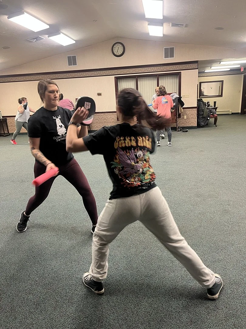 Two students training in a room with padded floor, one holding a pink dumbbell and the other wearing a black t-shirt with a graphic, engaged in a fitness exercise with other people working out in the background.
