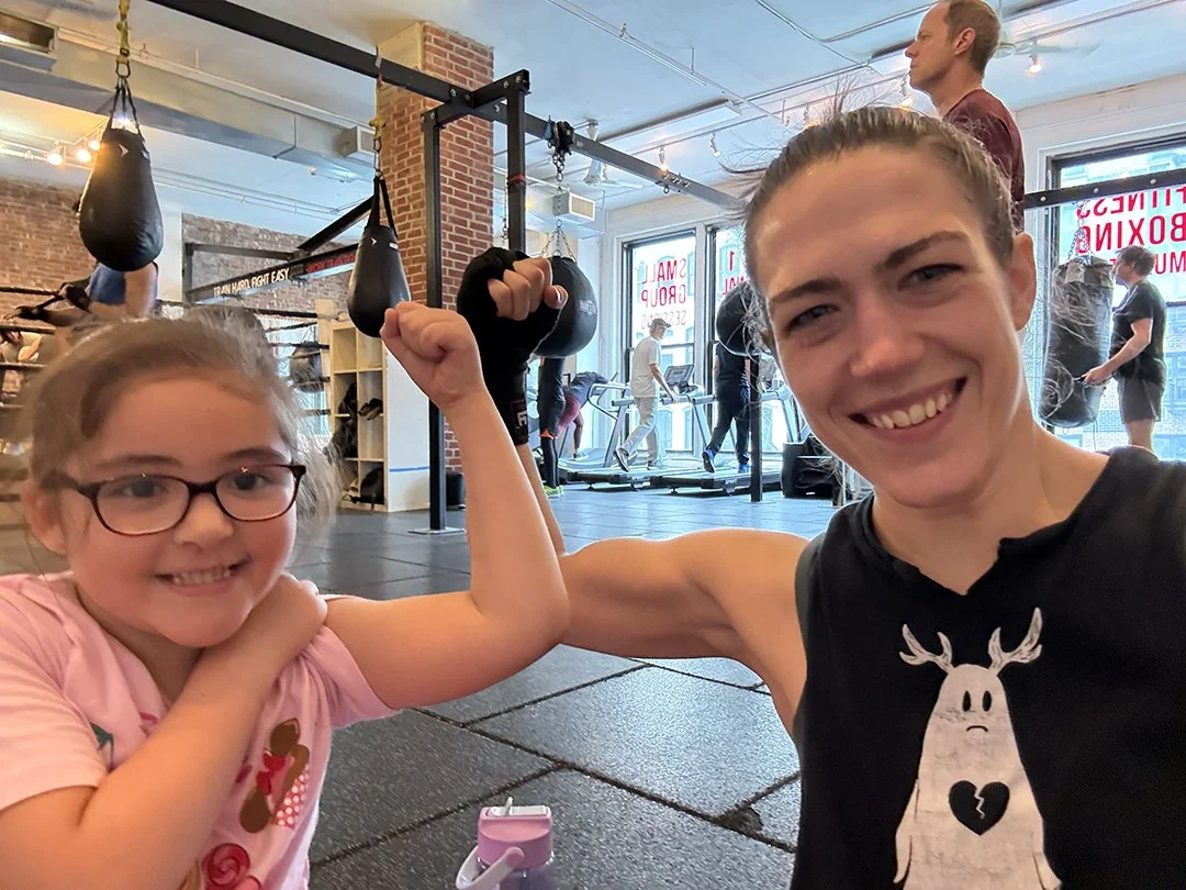 High Kick's instructor, Rae Switlick, poses with one of her young trainees, both flexing their muscles in the gym and smiling at the camera. The gym has brick walls, punching bags, and people working out in the background.