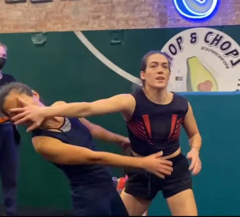Two women involved in a physical altercation inside a gym, with gym equipment and a logo in the background.