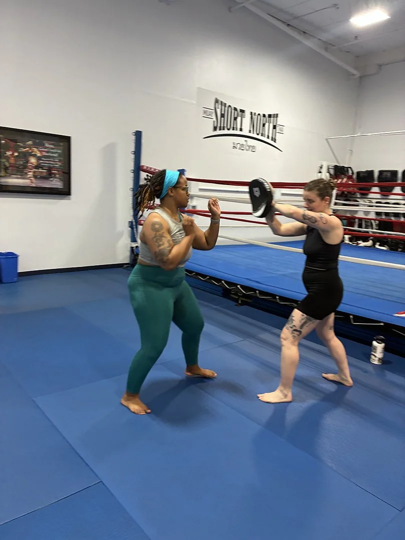 Two women training in a boxing gym, one holding focus mitts and the other punching with gloves, on a blue mat in front of a boxing ring.