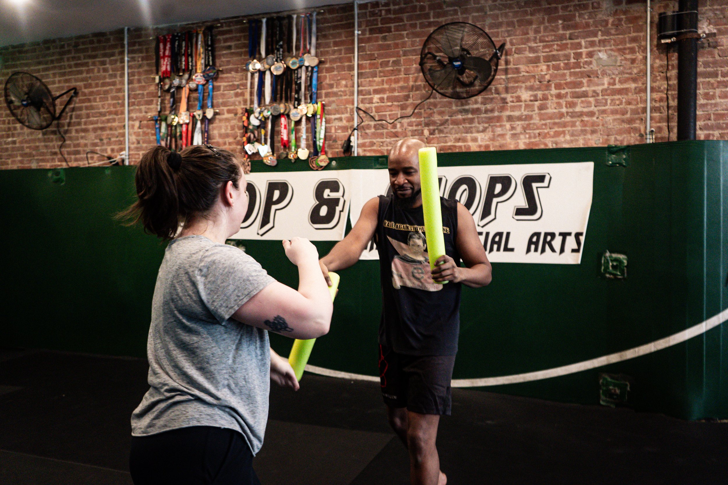 Two students are practicing martial arts with foam sticks at the Chop and Chops MMA gym in Brooklyn. In the background, medals and ribbons are hanging on the brick wall.