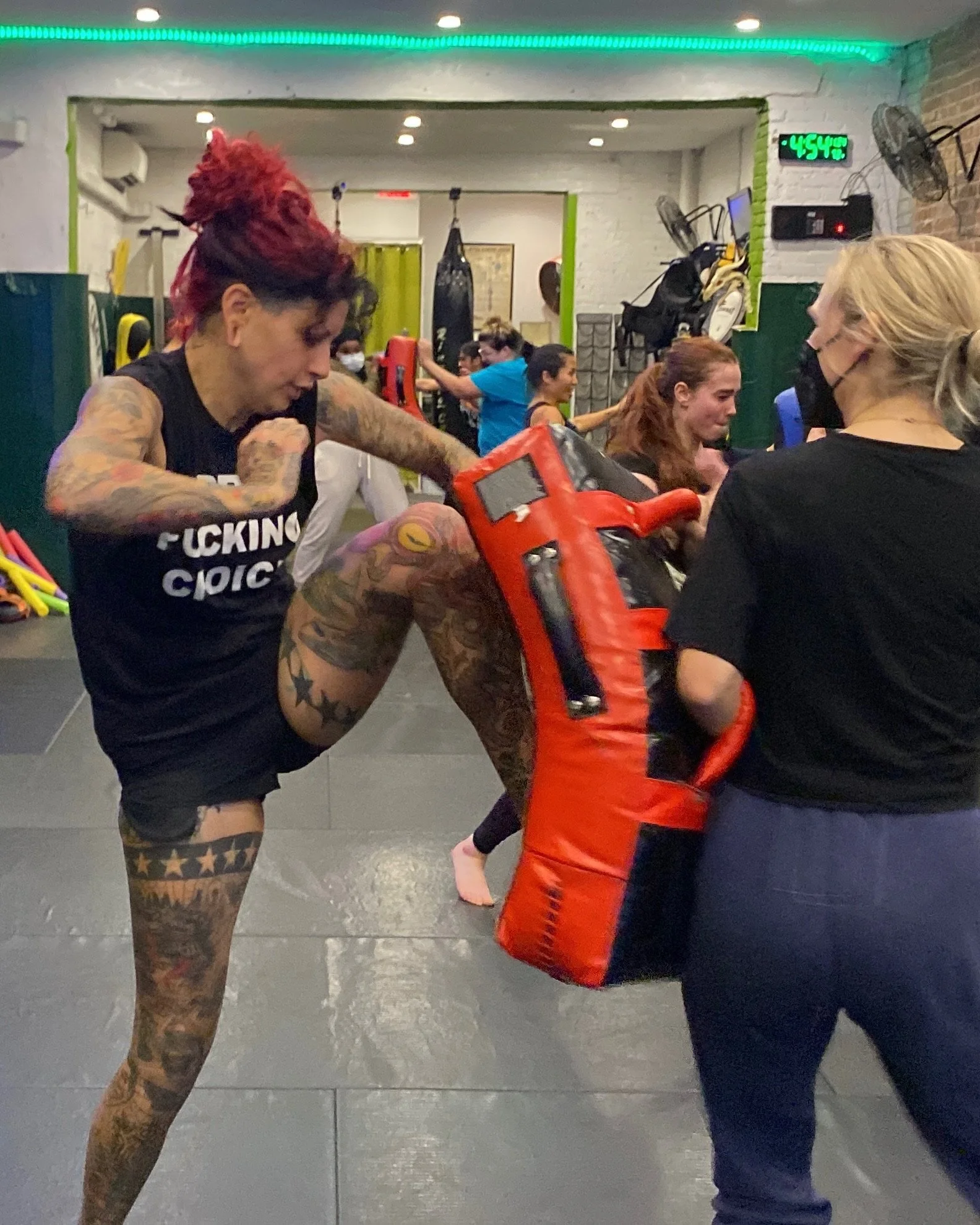 Boxing gym with people practicing boxing. Woman with red hair and tattoos wearing boxing gear, including red gloves and a black tank top. Other individuals in the background adjusting boxing equipment. Digital clock showing 4:54.