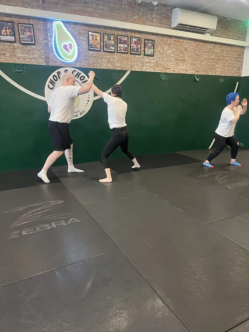 Two people practicing martial arts with a trainer in a gym. The instructor is giving a high five to a woman, while another woman with blue hair is preparing for her turn.