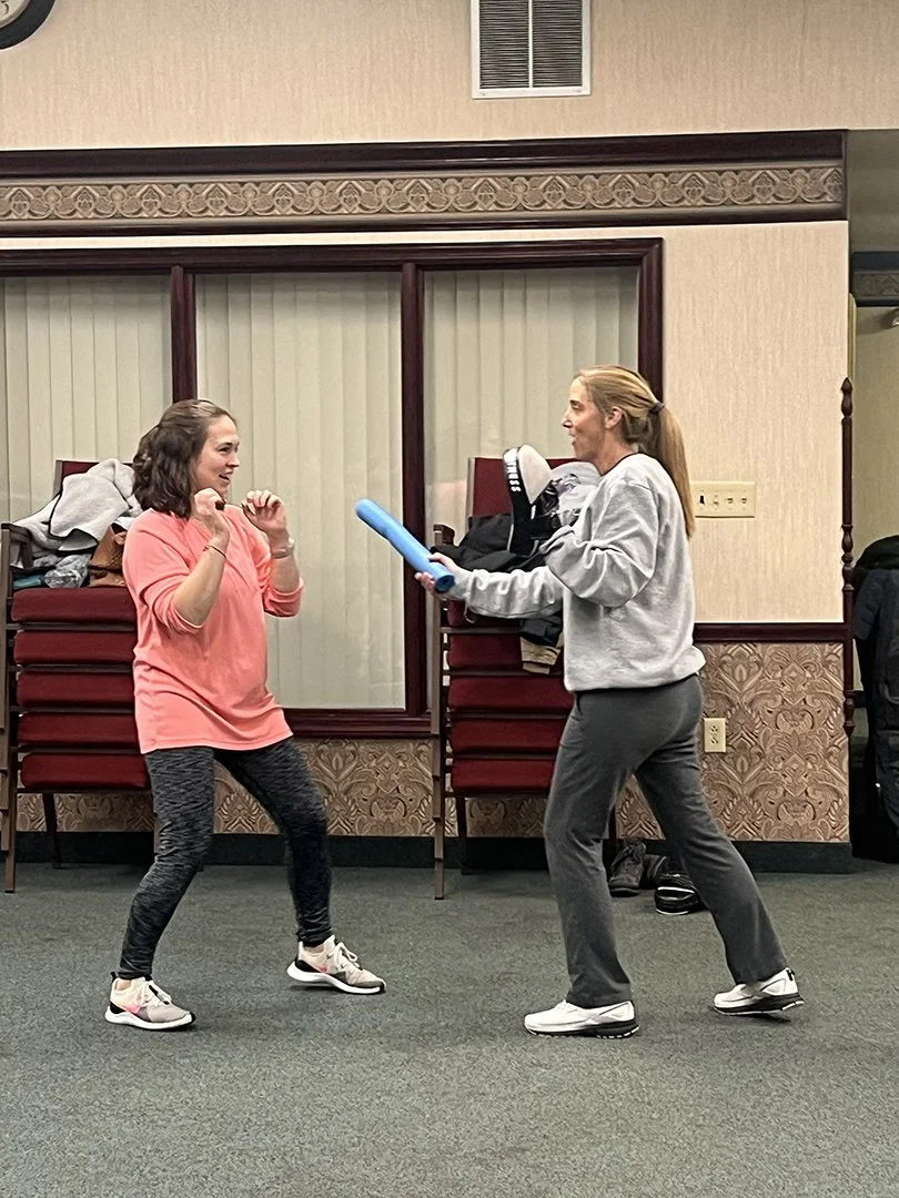 Two students practicing boxing indoors, one in a pink shirt and black leggings, the other in a gray hoodie and gray sweatpants, with foam noodles.
