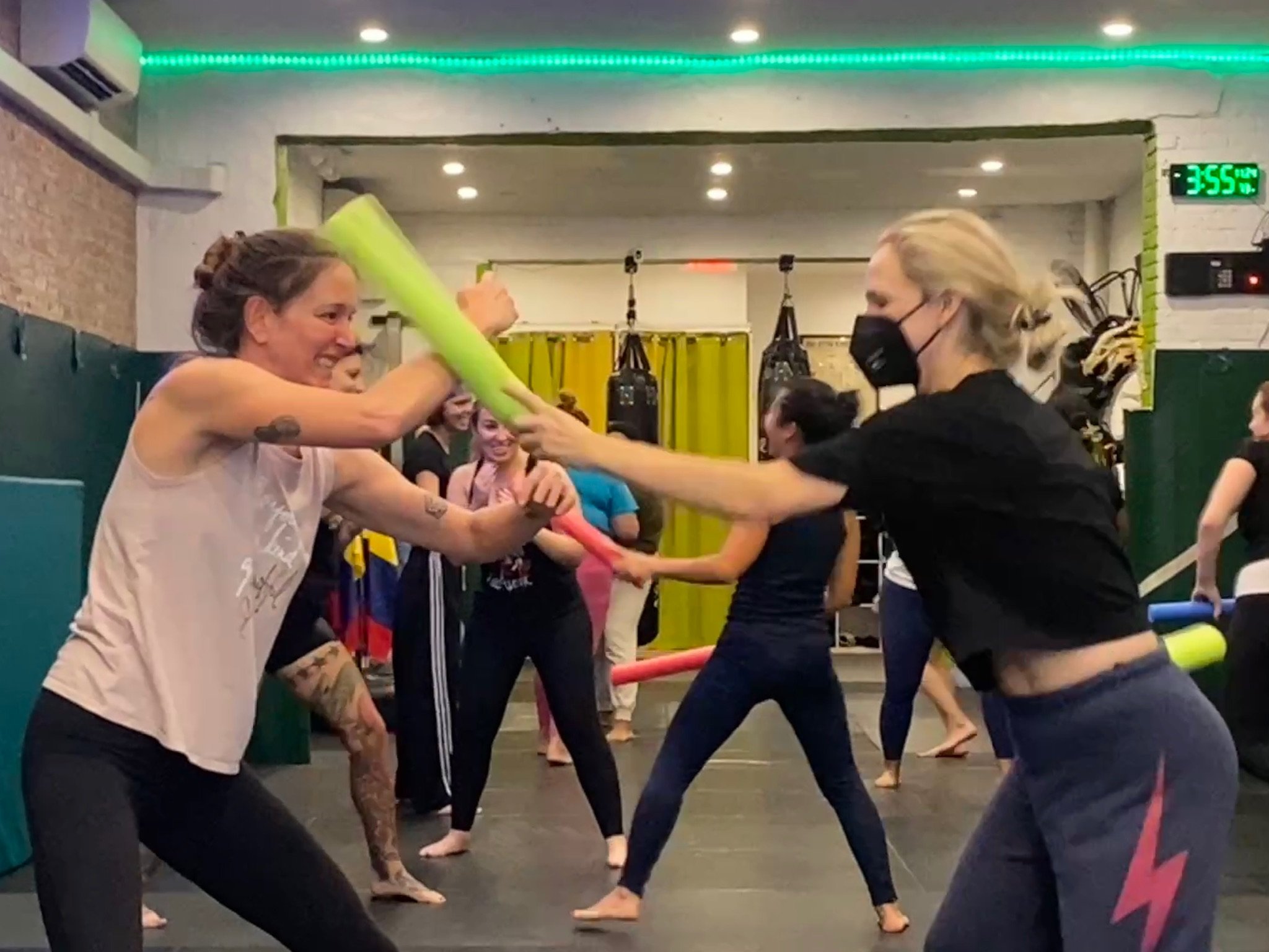 People participating in a fitness class at a gym, engaged in a workout with foam noodles, some wearing workout clothes and masks, in a brightly lit room with a digital clock showing 3:55.
