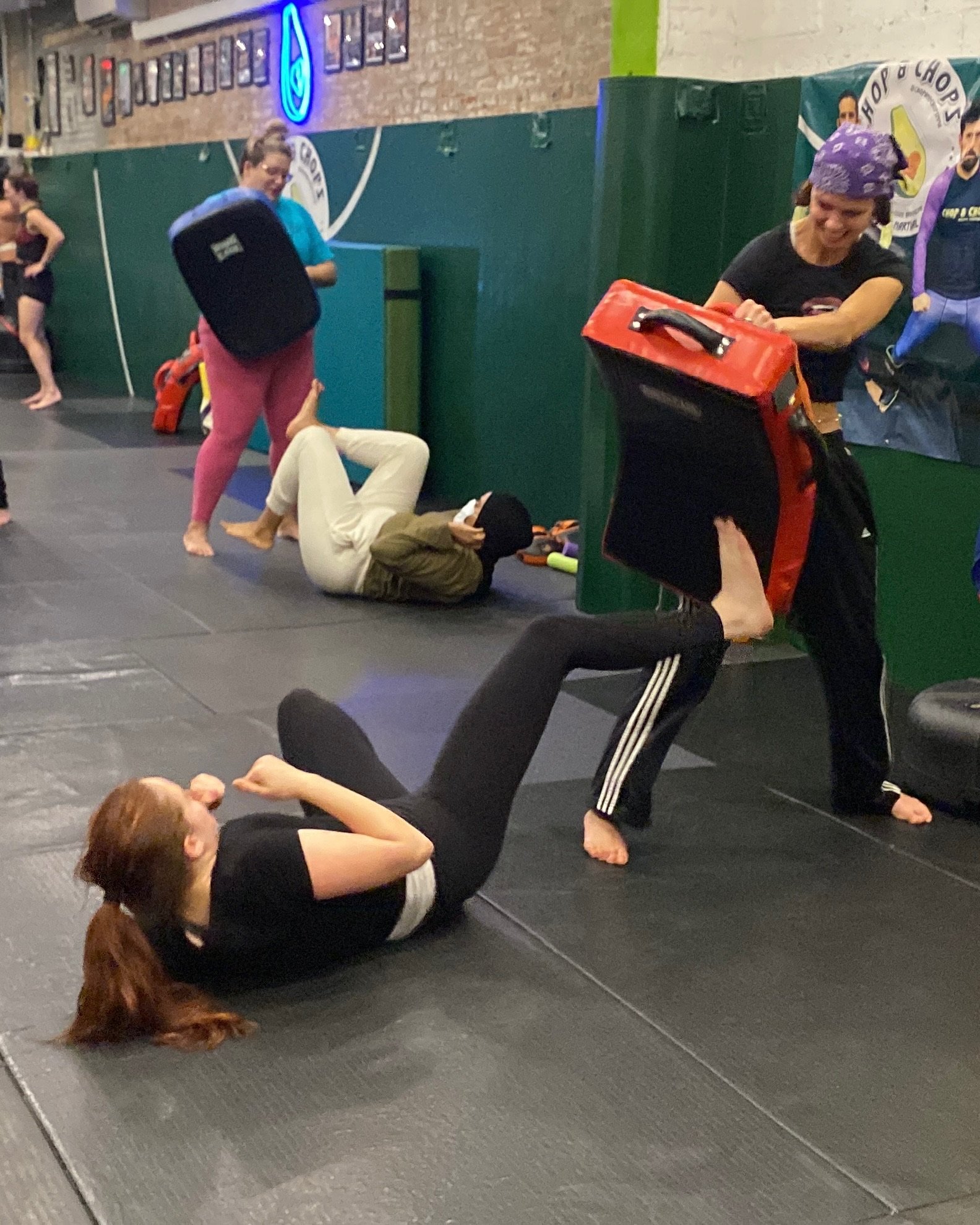 People in a martial arts gym practicing joint locks and grappling techniques on mats, with some lying on the floor and others standing, in a training session.