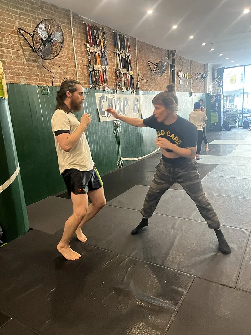 Two women practicing self-defense or martial arts in a gym. One woman is wearing a black T-shirt and camo pants, and the other is wearing a white T-shirt and black shorts. They are on a black padded mat, with medals hanging on the brick wall behind t
