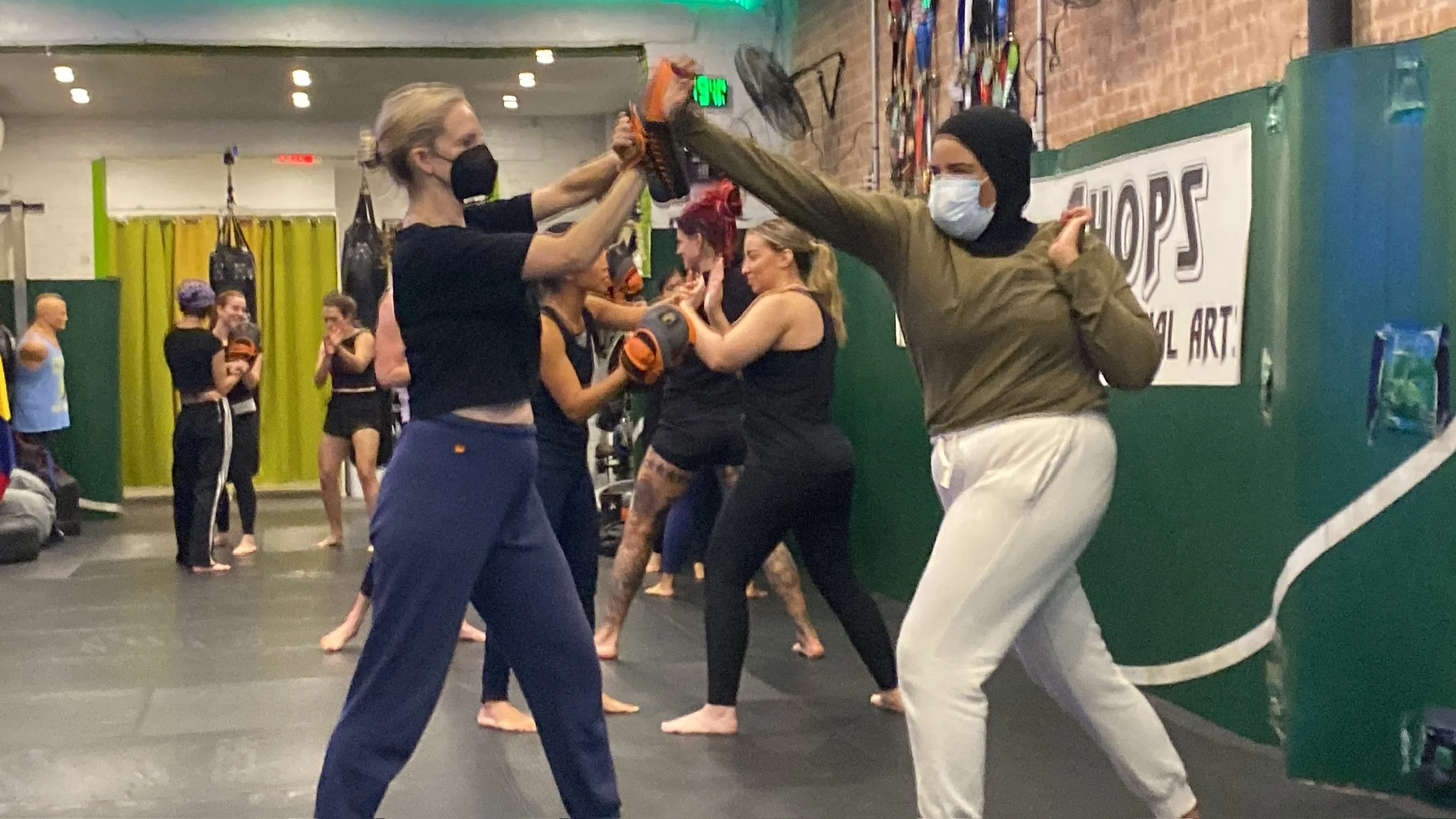 People practicing martial arts or self-defense in a gym or training studio, with some participants wearing masks and gloves, engaged in training exercises or drills.