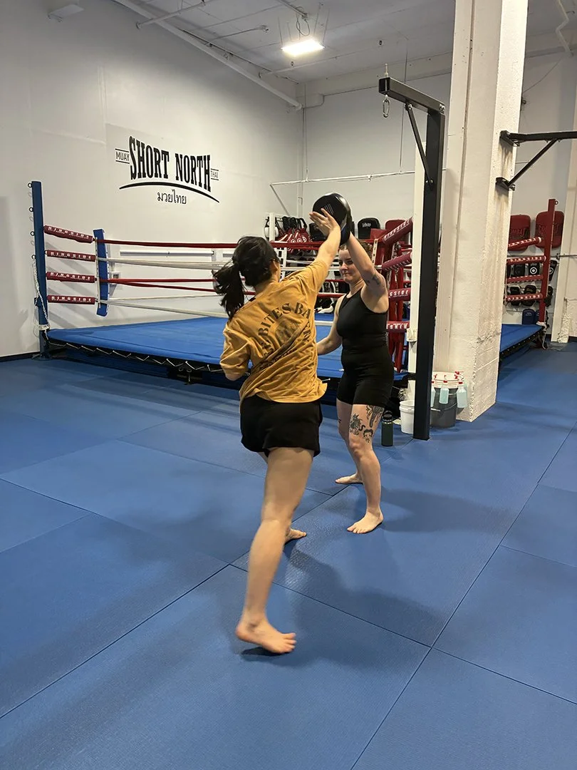 Two women practicing Muay Thai in a boxing gym. One woman in yellow jacket and black shorts is throwing a punch, while the other woman in black tank top and black shorts is holding a black pad, with a boxing ring and gym equipment in the background.