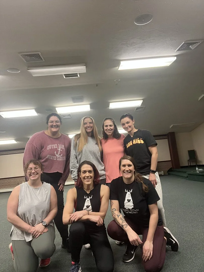 A group of seven women in a room with a green carpet and ceiling lights, posing for a photo. Some are wearing black shirts with a reindeer logo, others in casual clothes.