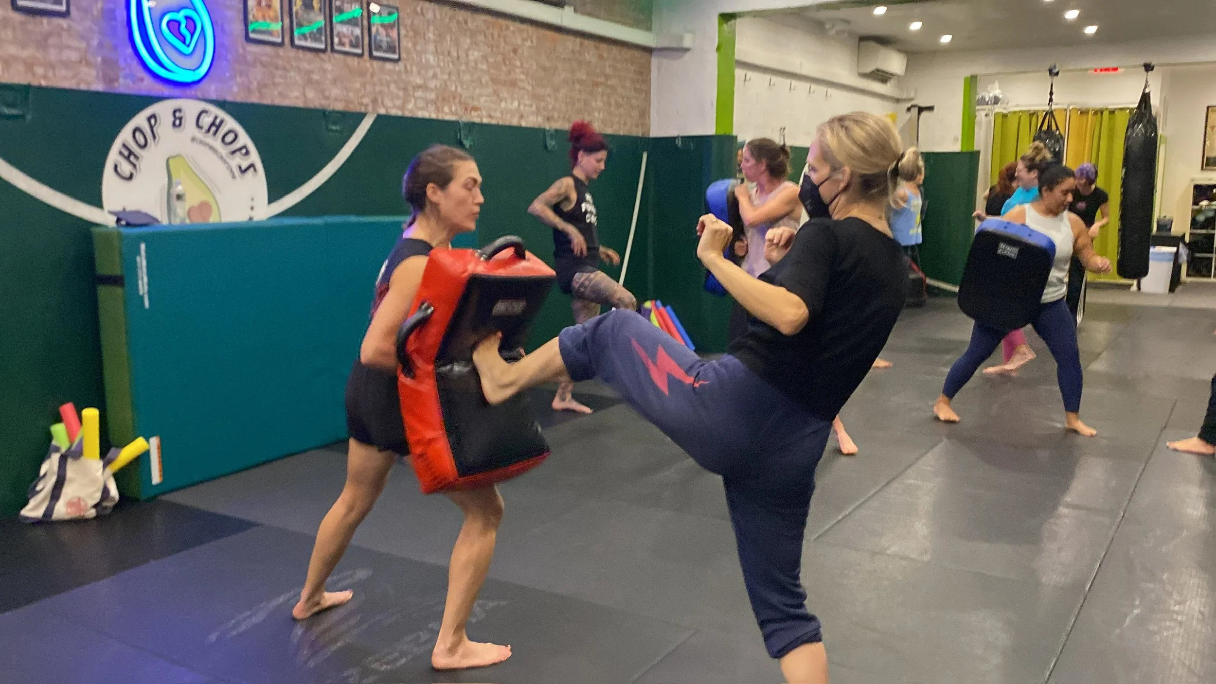 Women practicing martial arts in a gym, wearing fitness attire and masks, with some holding training pads and others engaging in sparring.