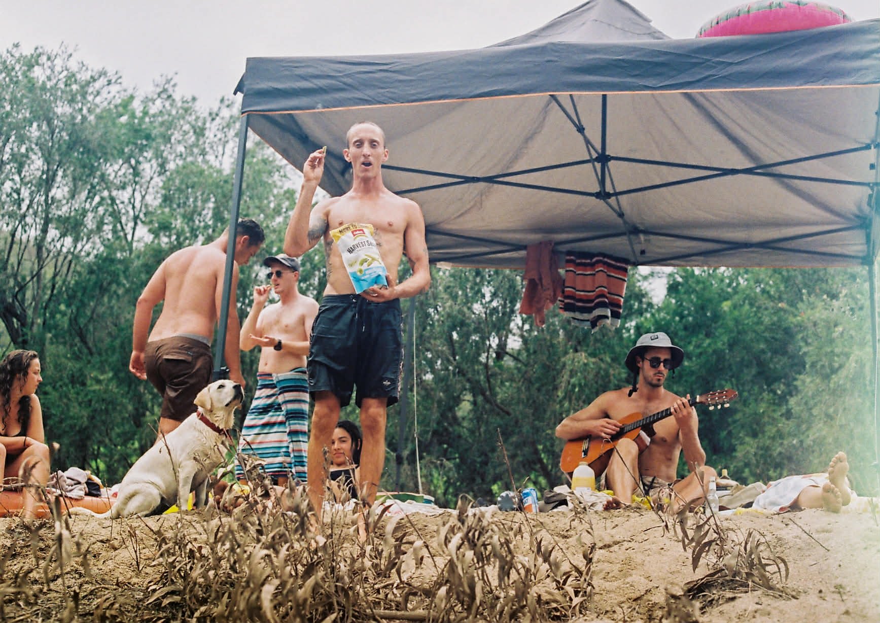 A group of shirtless people on a sandy beach with a canopy, one is sitting with a guitar, others are standing or sitting, with trees in the background. One person is holding a snack bag.