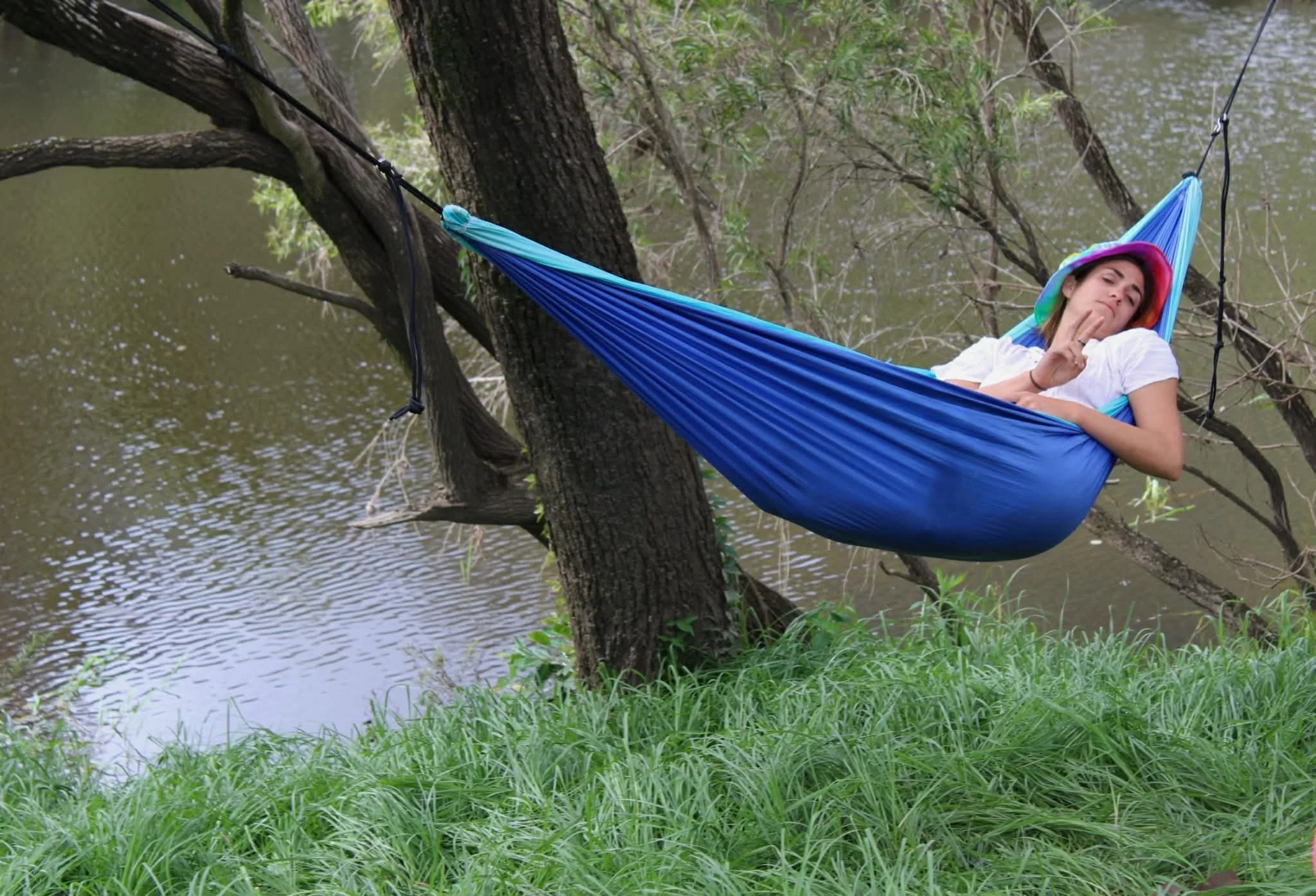 A woman lying in a blue hammock tied between two trees near a river, making a peace sign with her fingers and resting her head on a colorful hat, in a natural outdoor setting with green grass and trees.