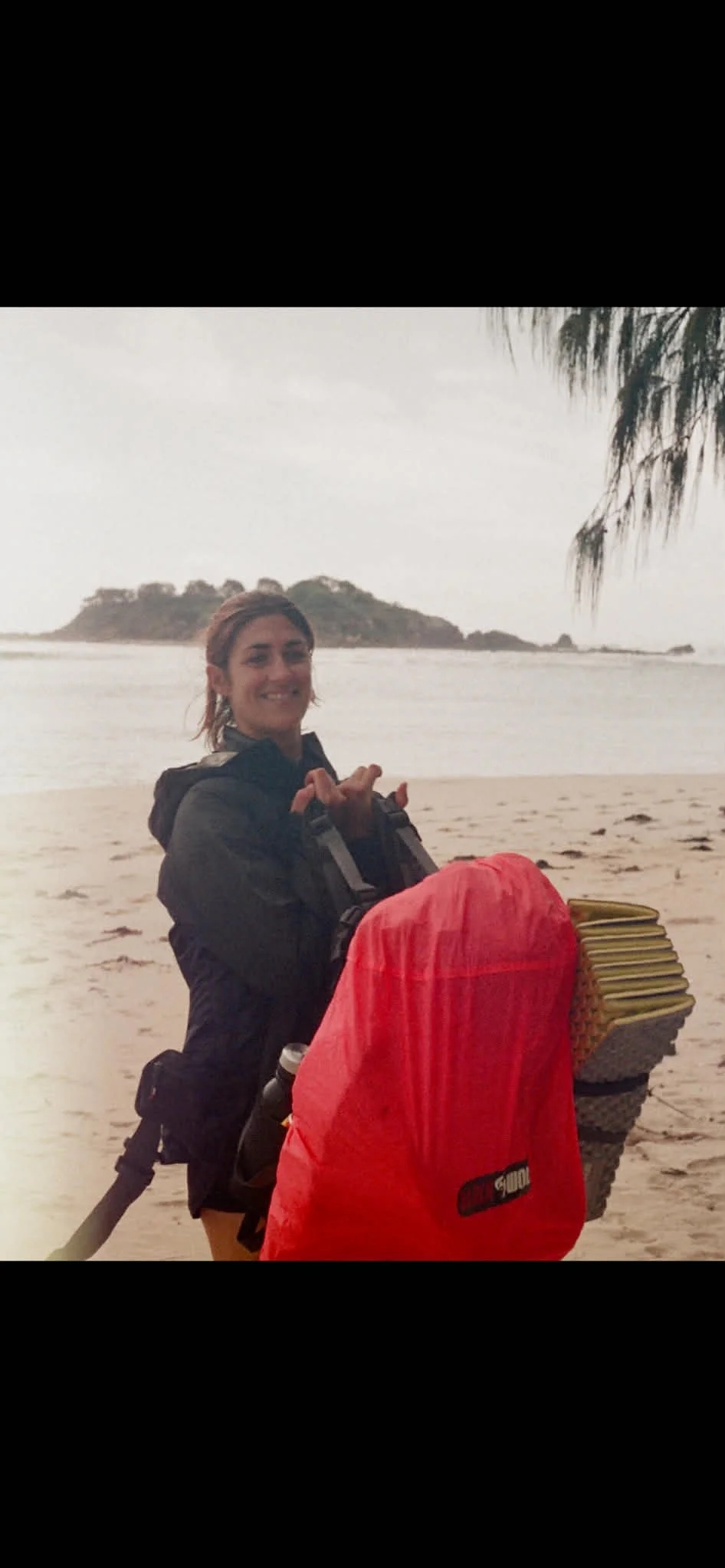 A woman stands on a beach with an island in the background. She is smiling, wearing a black jacket, and has a red backpack with yellow poles or objects attached.
