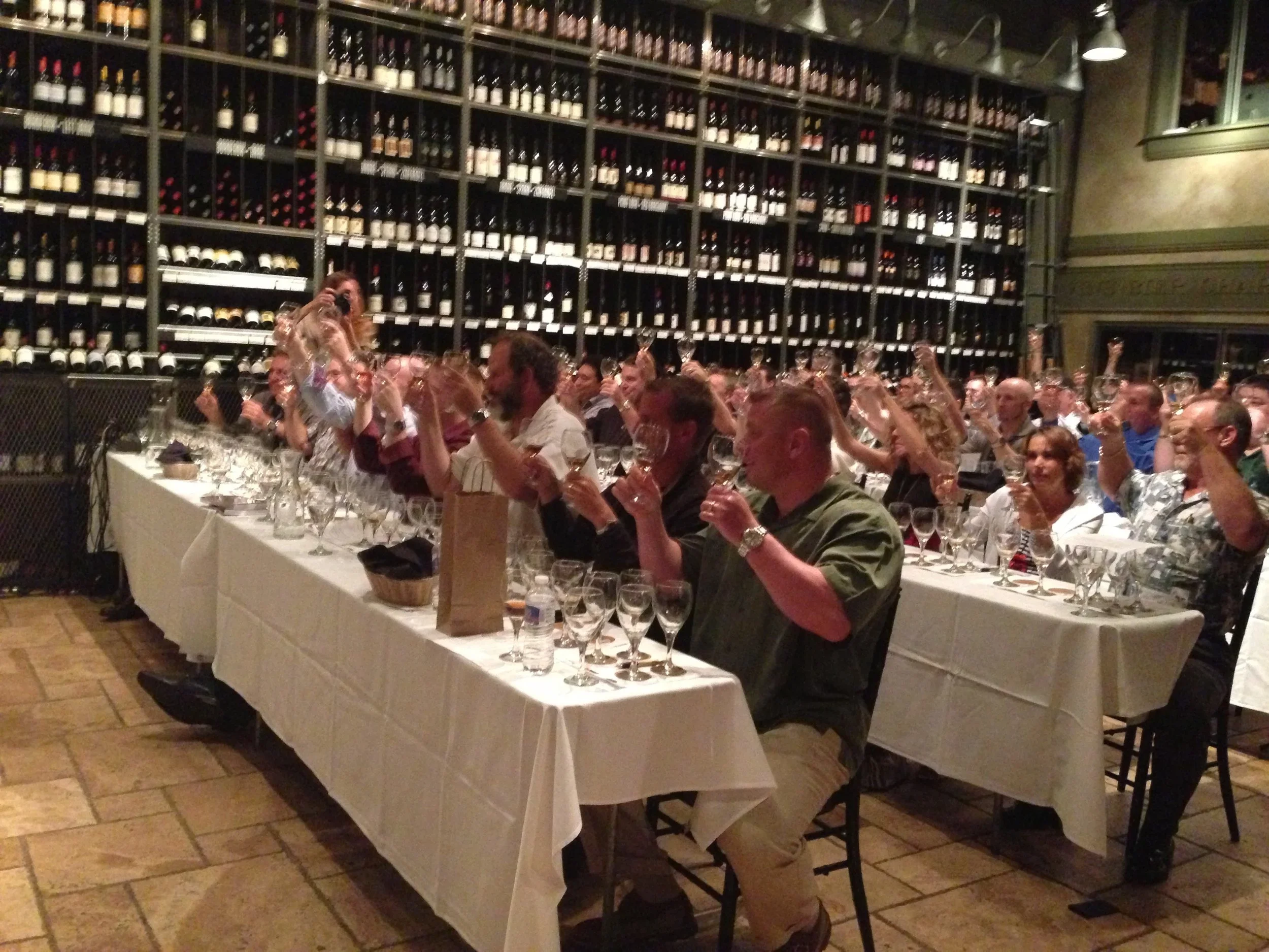A group of people sitting at a long table in a wine cellar, holding up wine glasses, possibly at a wine tasting or celebration event.