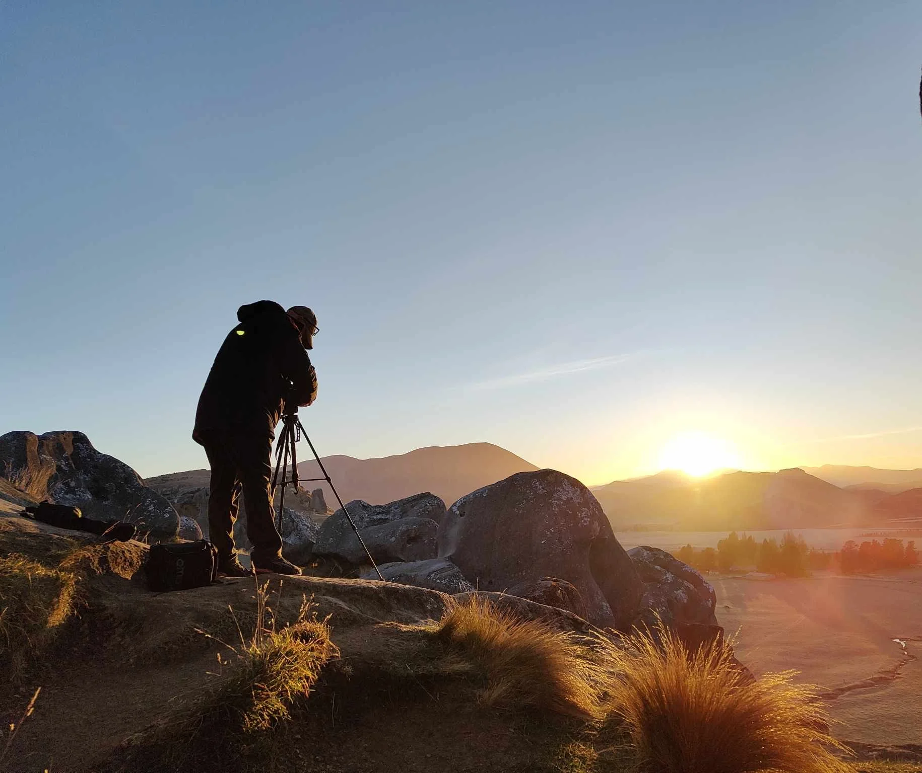 Photographer with tripod capturing a sunrise over scenic landscape with rocks and hills.