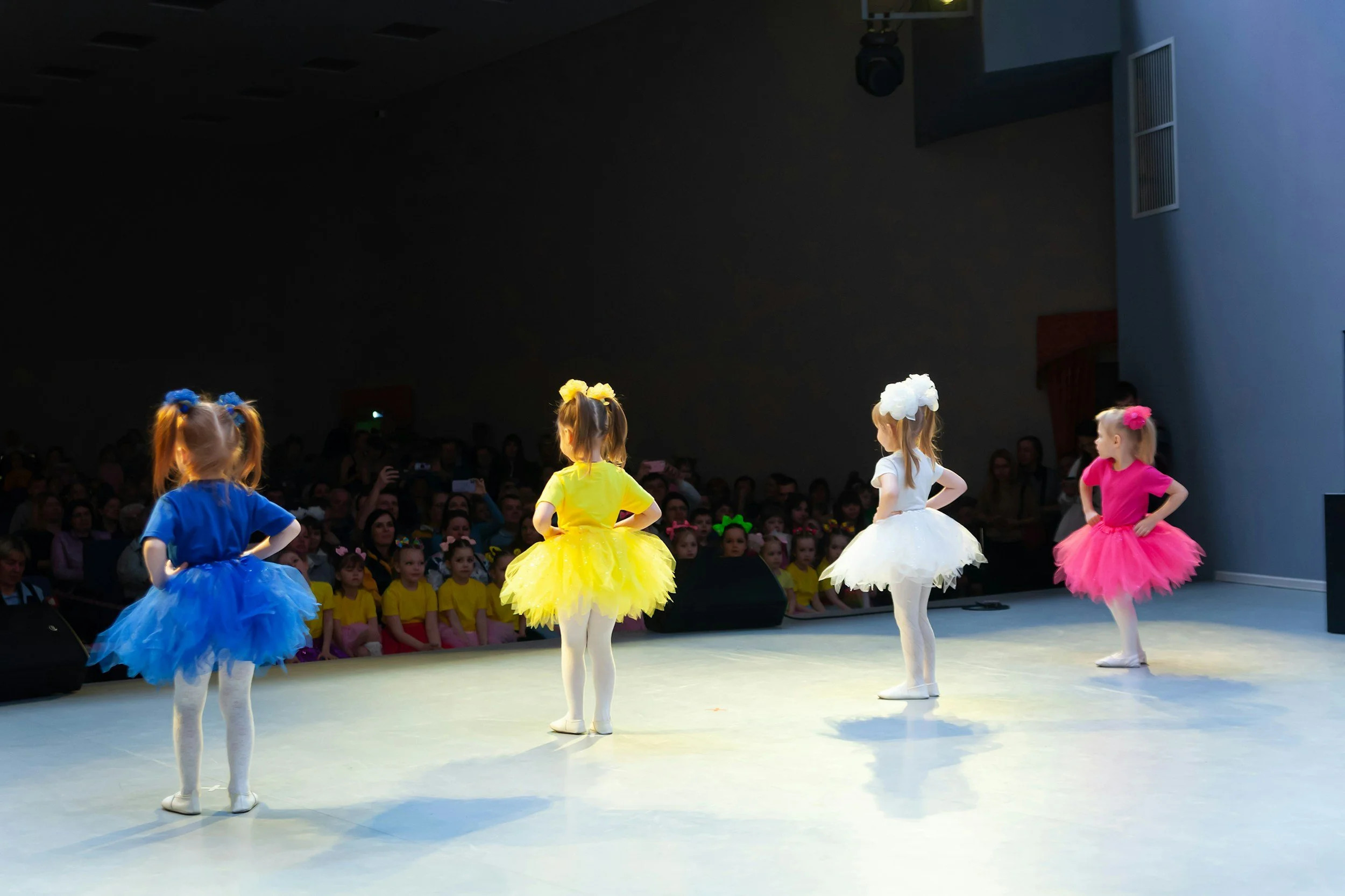 Four young girls in colorful tutus performing a dance on stage with an audience watching in the background.