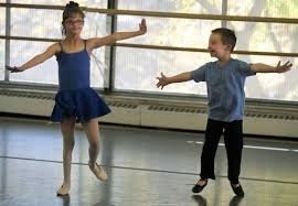 A young autistic girl in a blue ballet outfit and a young adhd boy in a grey shirt and dark pants dancing in a dance studio with a wooden floor and ballet barres.