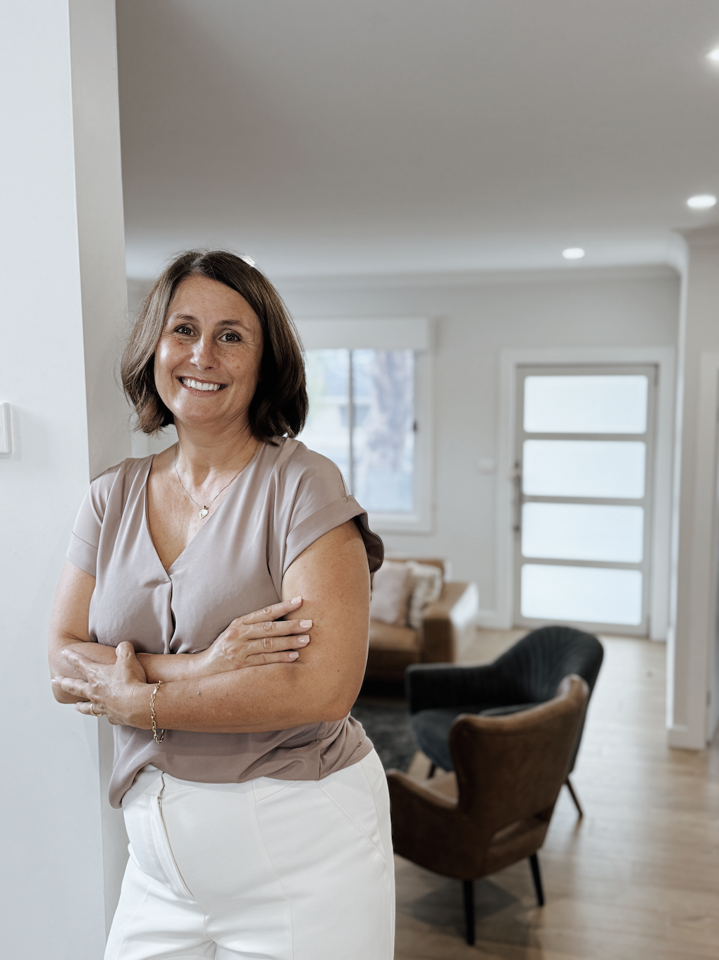 Tania, wearing a beige blouse and white pants standing in a modern living room with natural light, a sofa, and chairs.