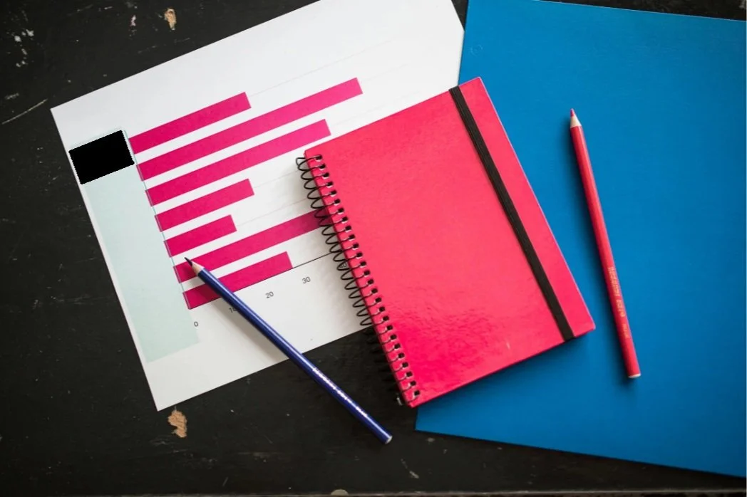A desk with a printed chart, a pink spiral notebook, a blue folder, and two red pens.