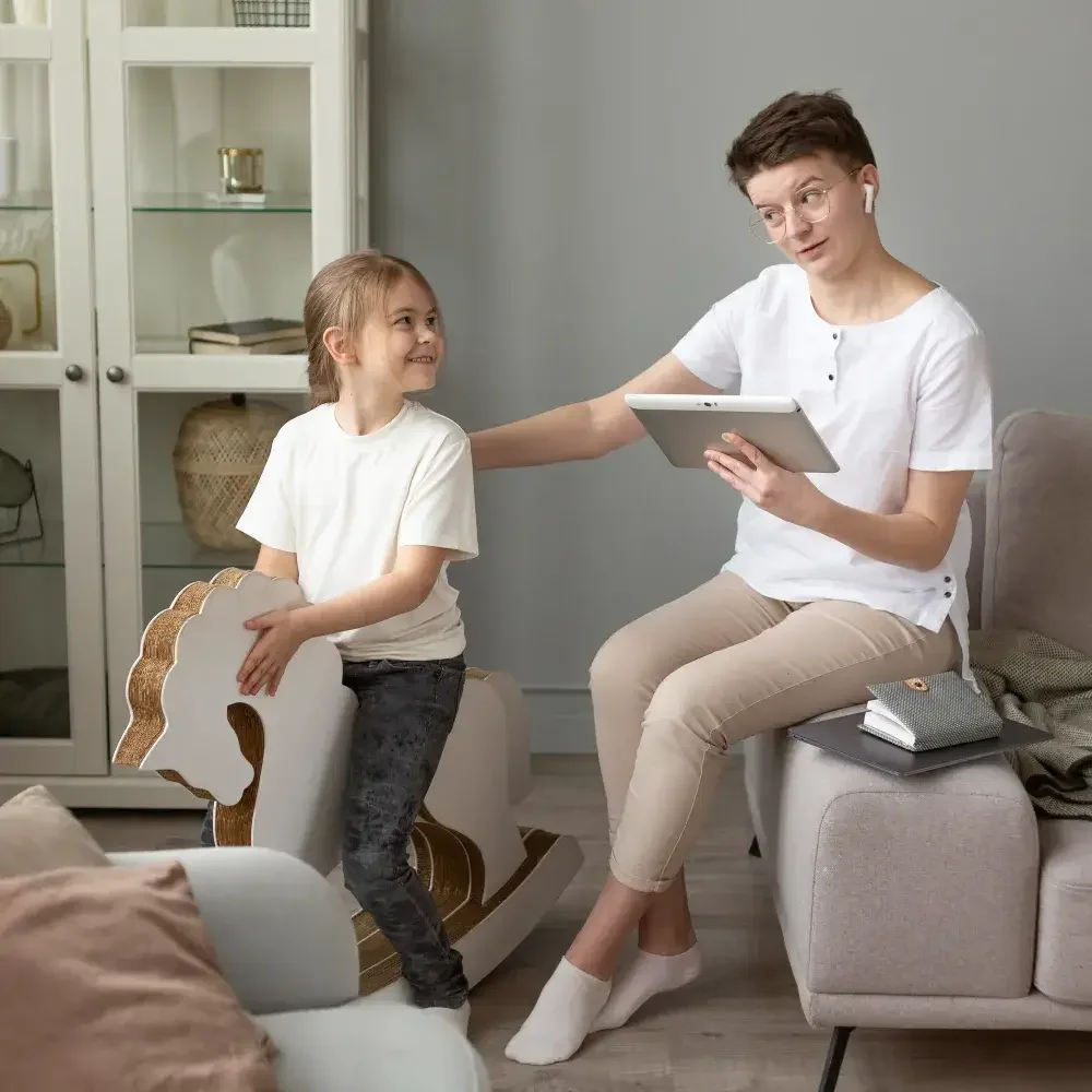 A woman and a young girl in a living room, smiling and talking. The woman is sitting on a couch with a tablet in her hand, while the girl is holding a white cutout of a horse. The room has a gray wall, a glass-front cabinet, and cozy furniture.