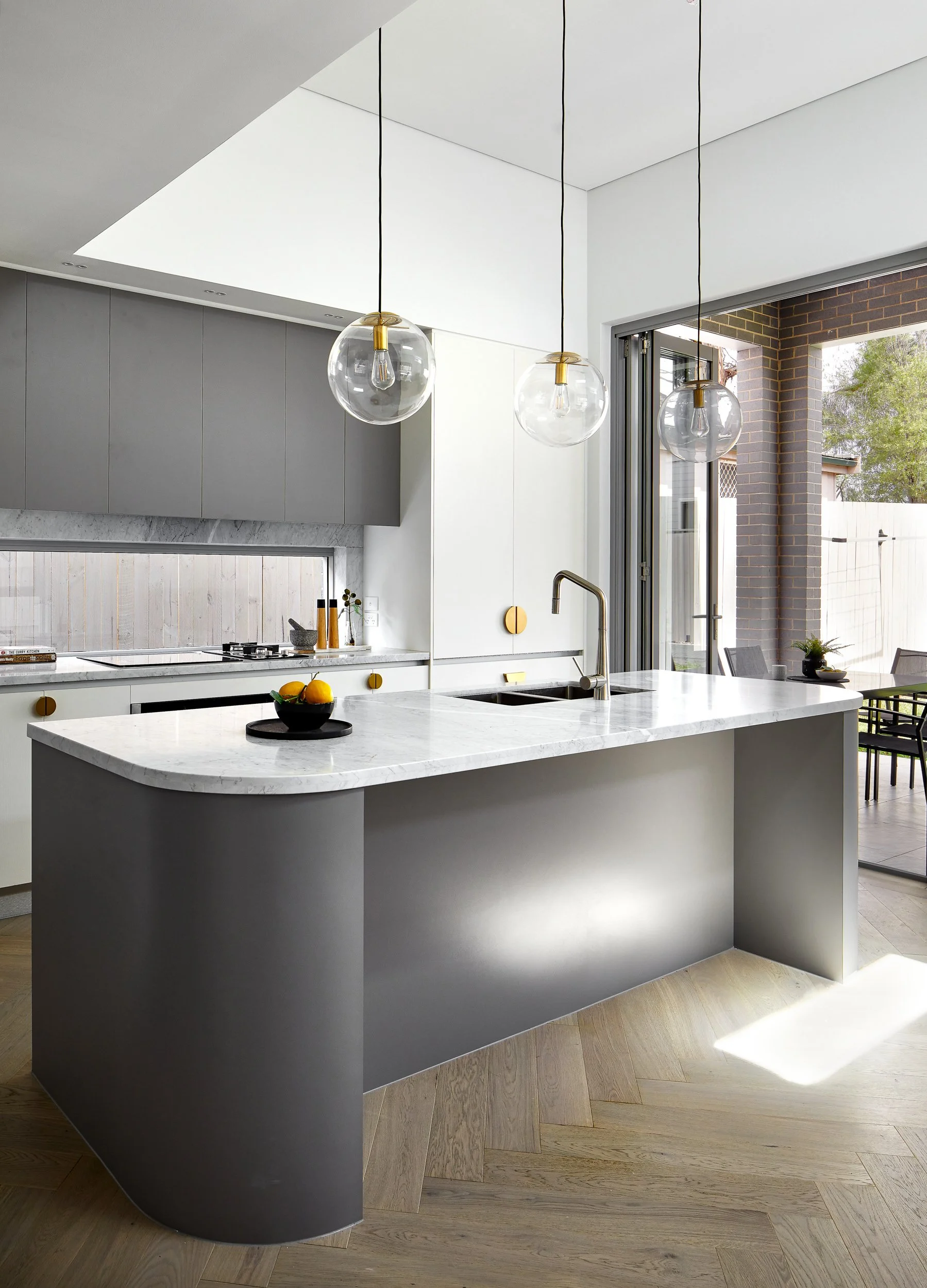 Modern kitchen with white marble countertops, gray cabinetry, hanging glass globe pendant lights, and a sliding glass door leading outside.