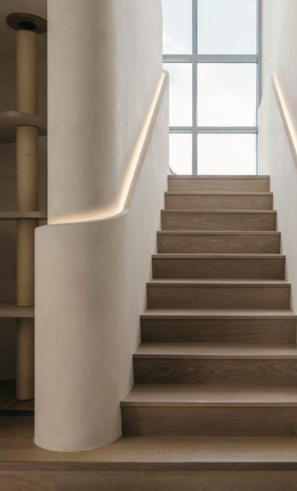 Wooden staircase with a white curved wall and handrail, large window at the top letting in natural light.