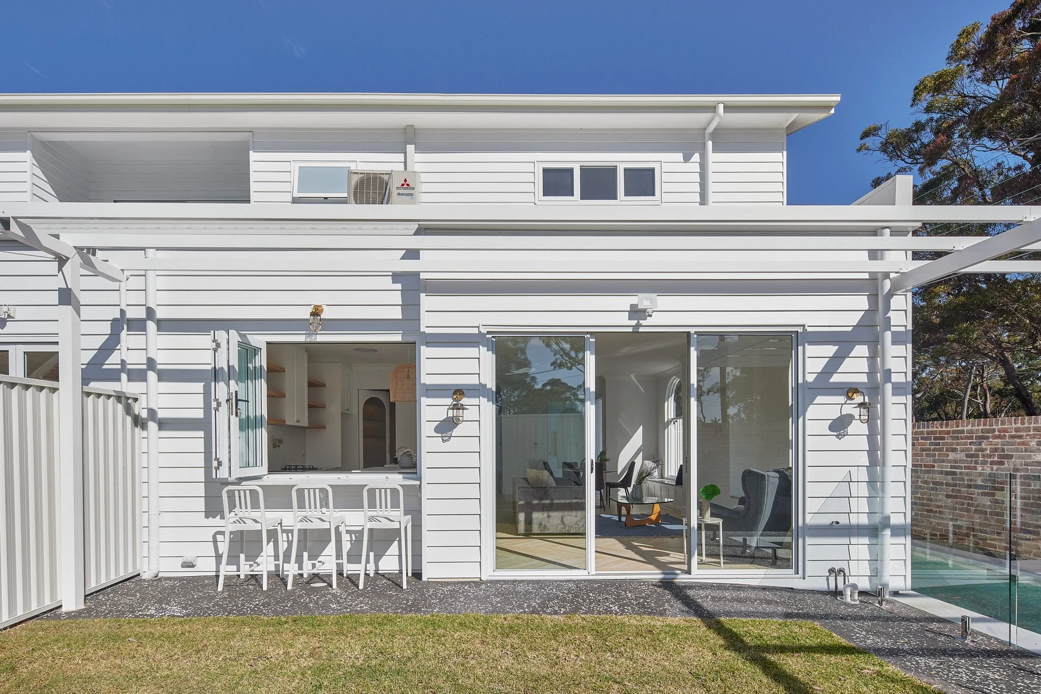 White two-story house with large sliding glass doors, a small outdoor bar with four white chairs, and a backyard with a lawn and a pool. Clear blue sky and trees in the background.