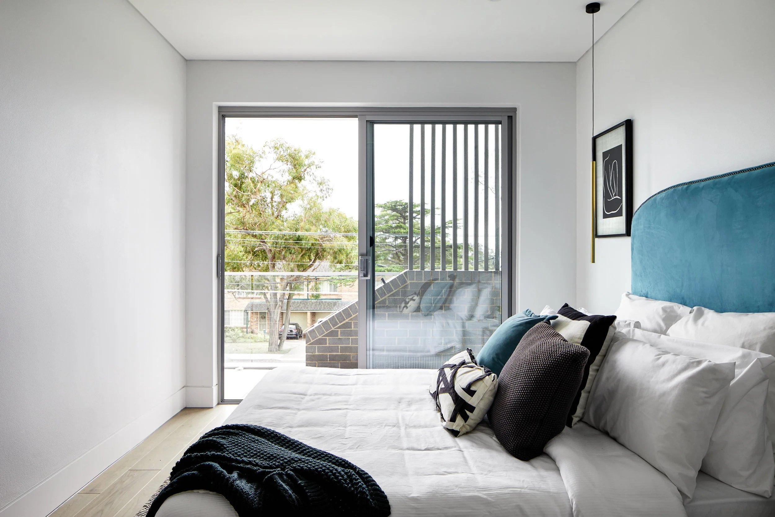 Modern bedroom with white walls, large glass sliding door leading to balcony, blue upholstered headboard, and bed with white linens and assorted pillows.