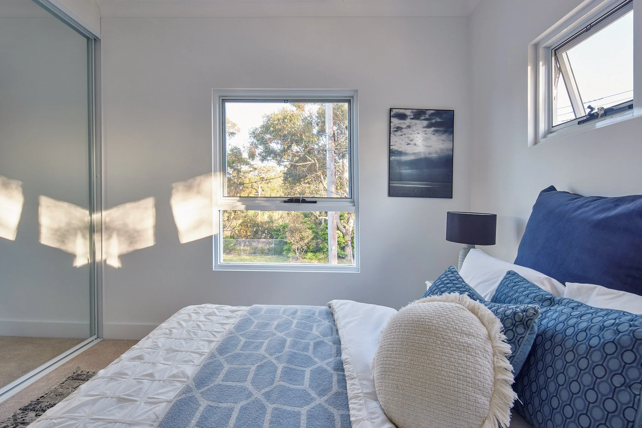 A modern bedroom with two windows, white walls, a bed with blue and white bedding, and a black bedside lamp. Sunlight creates shadows on the wall.