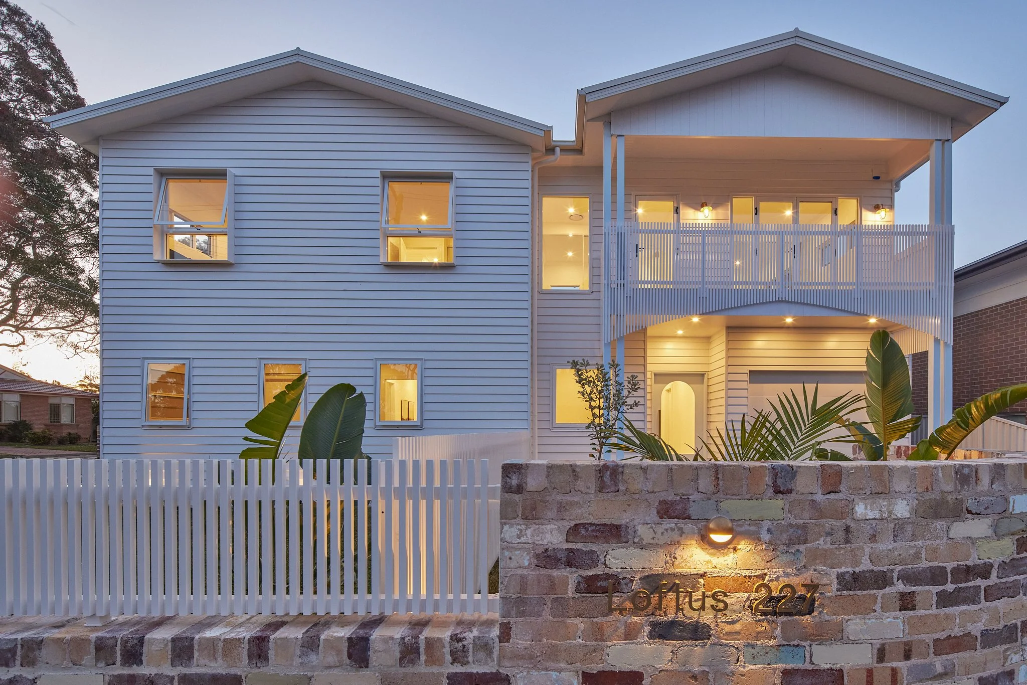 Two-story modern house with white siding and multiple windows, illuminated from inside, featuring a large balcony on the upper level and a garden area with tropical plants in front, enclosed by a brick and white picket fence with a small light and gate at the front.