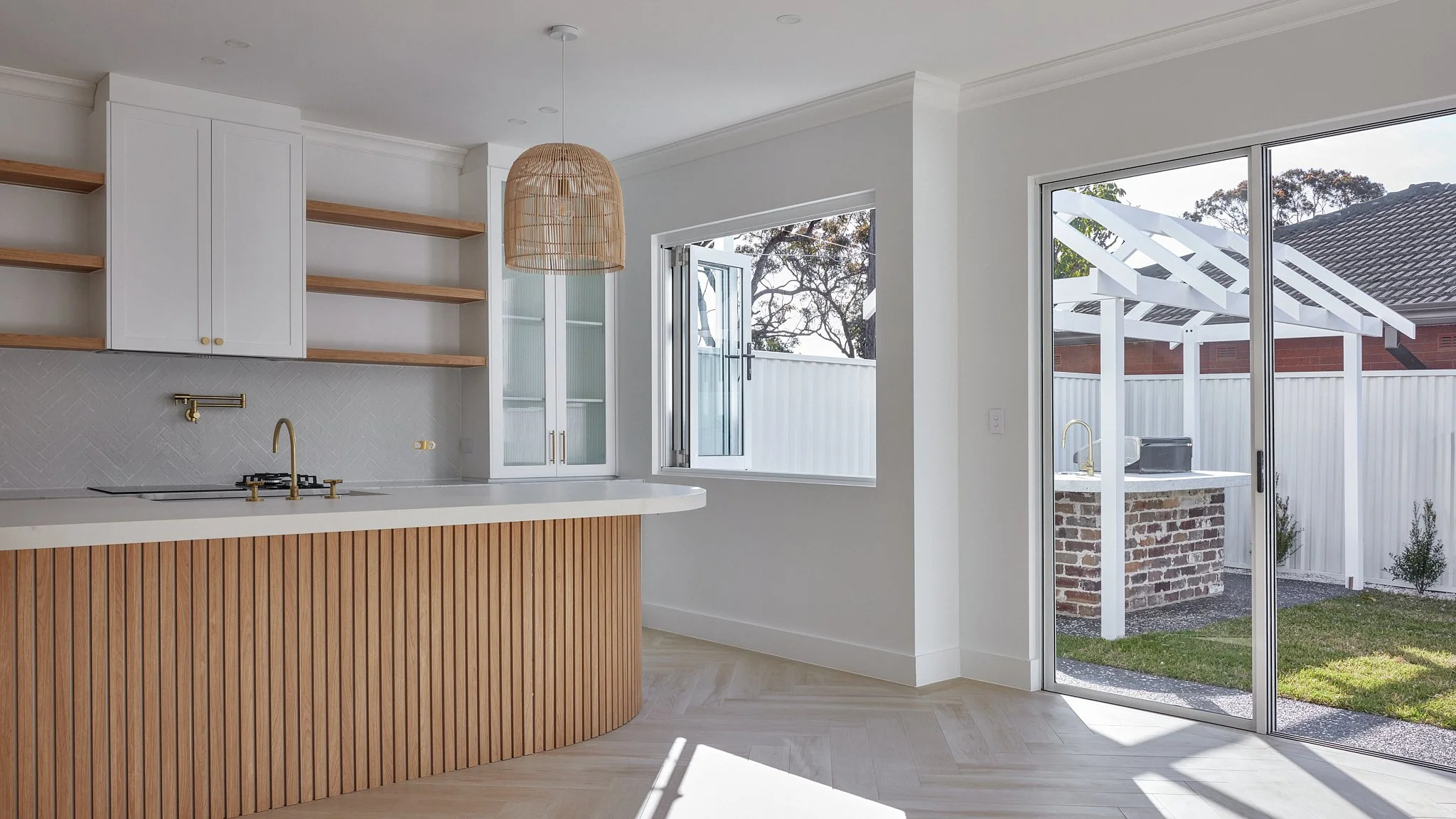 Modern kitchen with white cabinets, open shelves, a wooden island, a large window, and a sliding glass door leading to an outdoor area with a brick structure and pergola.