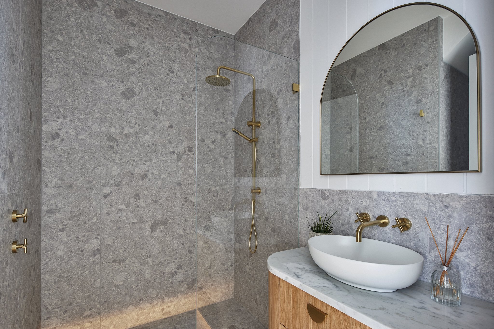 Modern bathroom featuring a walk-in shower with gold fixtures, a marble countertop with a vessel sink, a large arched mirror, and minimal decor.