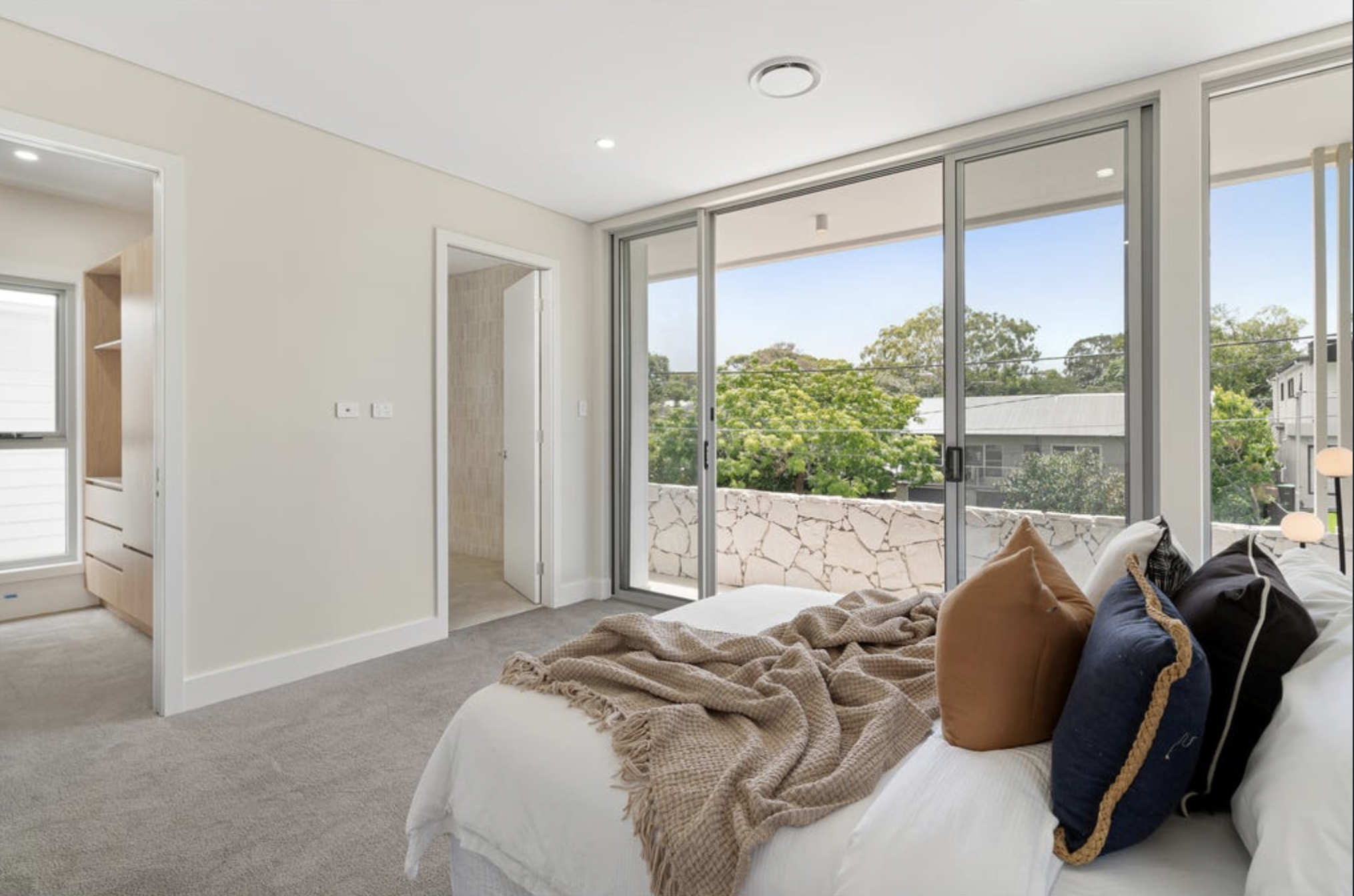 Modern bedroom with large sliding glass door leading to balcony, white walls, beige carpet, and a bed with combination of brown, black, and white pillows, and beige blanket.