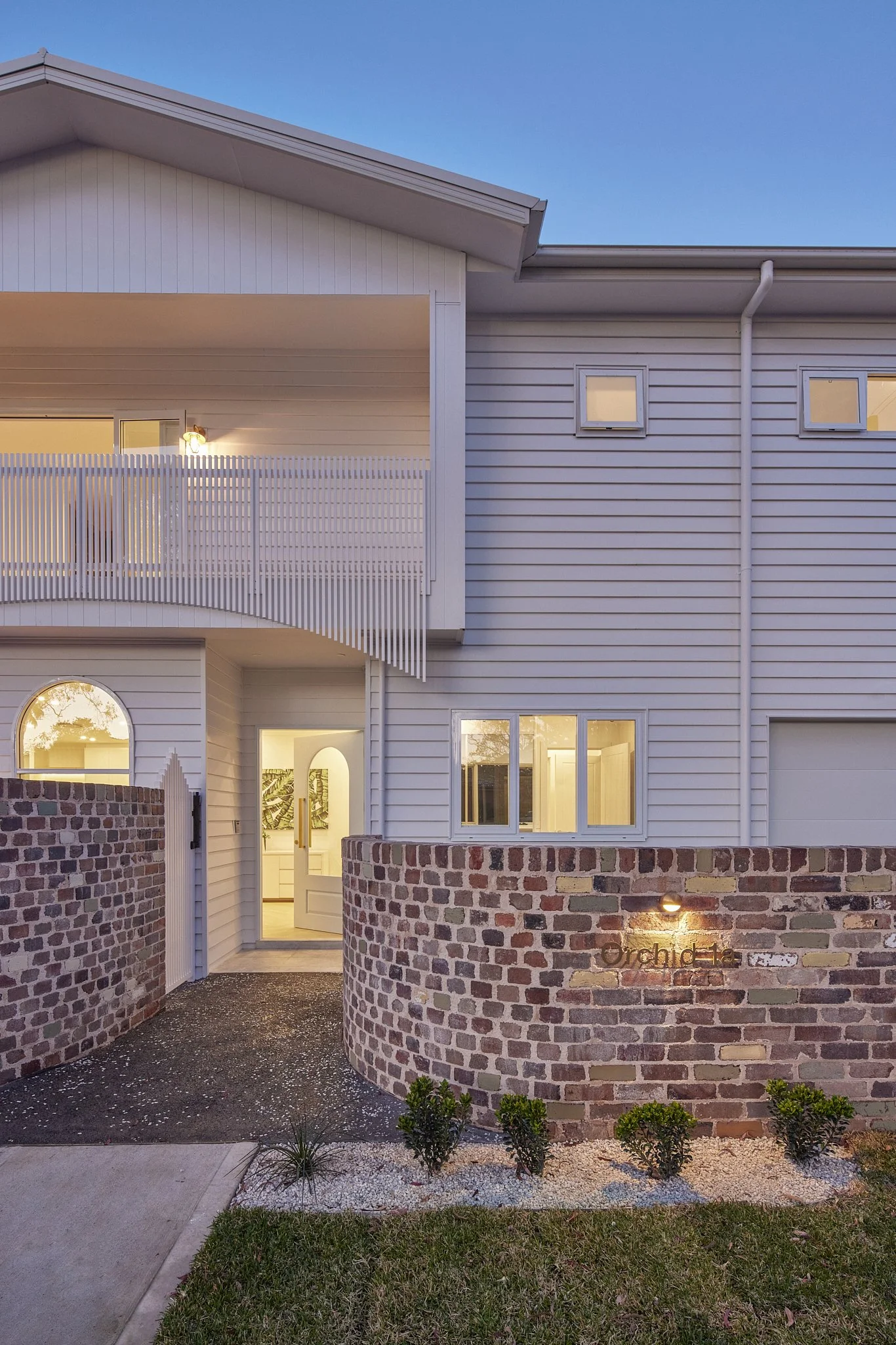Modern white house with brick curved wall and small garden, illuminated by exterior lighting, during dusk.