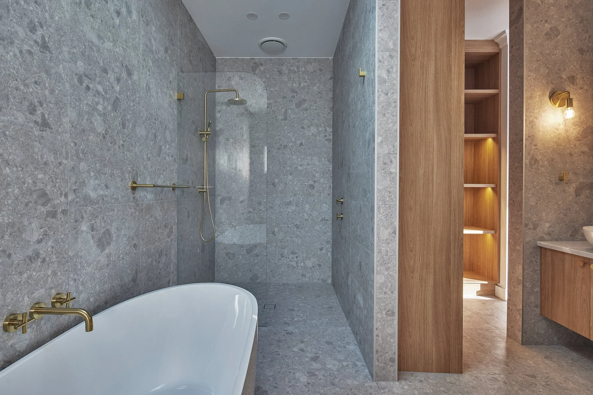 Modern bathroom with gray stone tiles, a white bathtub with brass fixtures, and a separate shower area with a brass showerhead. There is a wooden cabinet and a beige countertop with a sink, subtle lighting, and an open wooden shelving unit in a well-