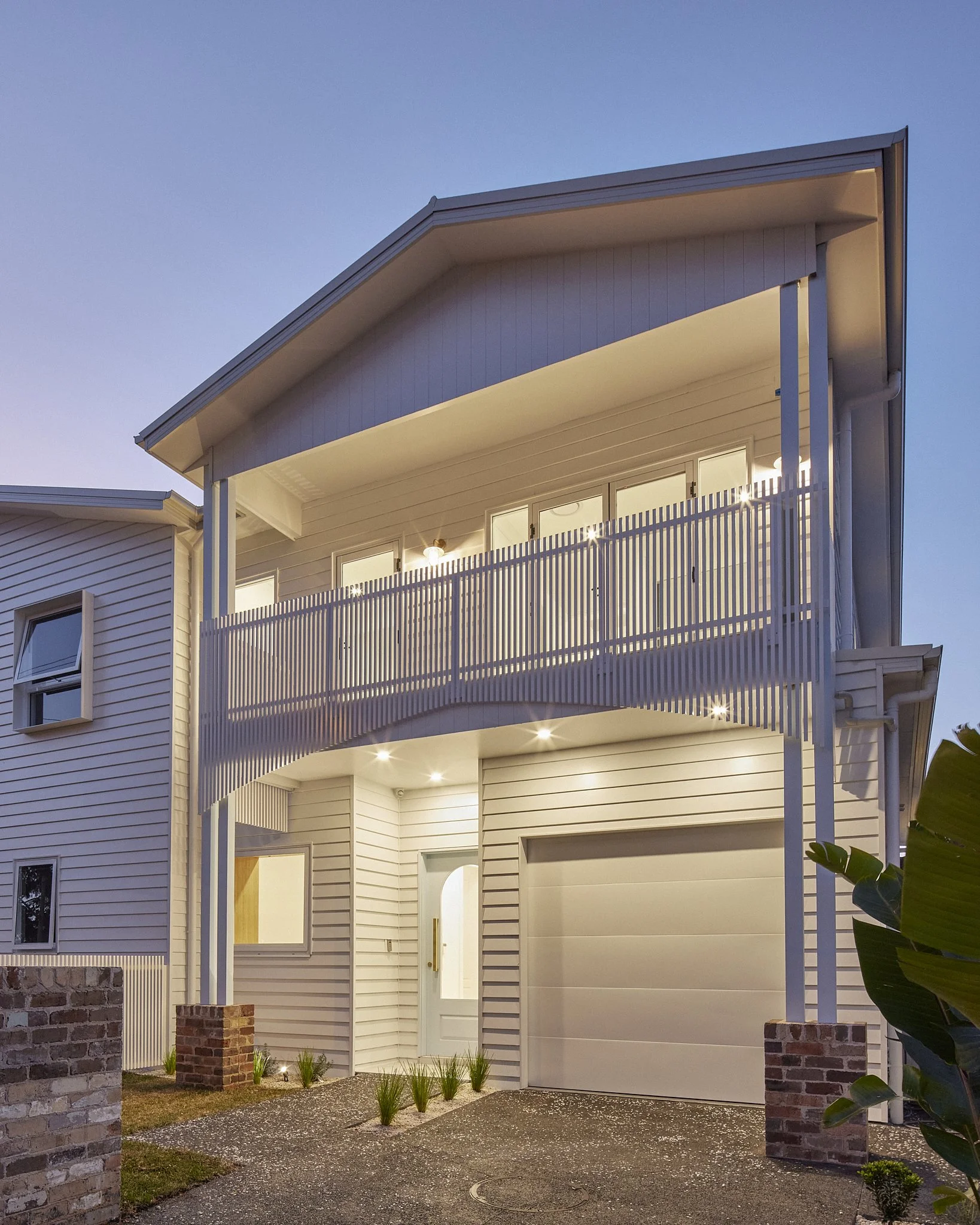 A modern two-story white house with a curved balcony, illuminated by outdoor lighting, at dusk.