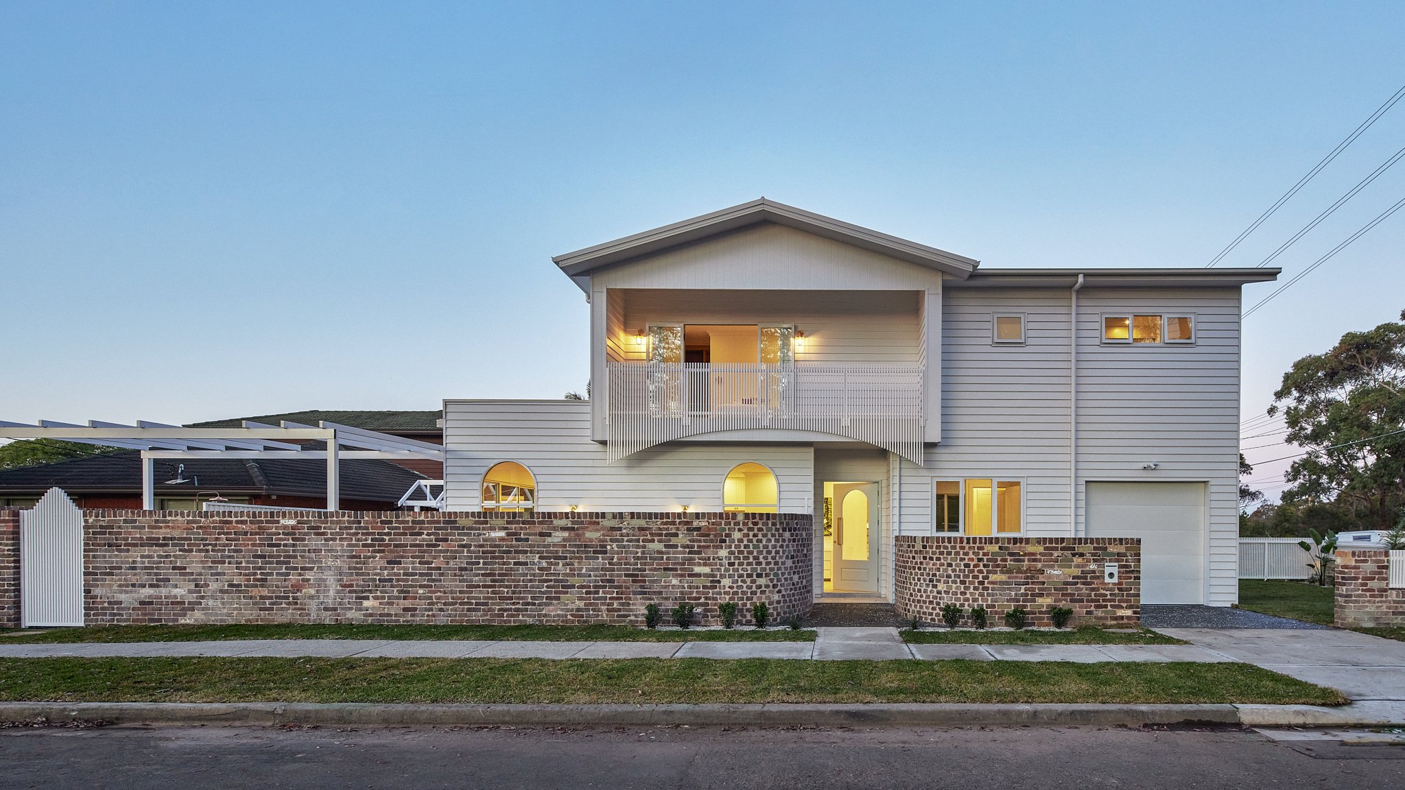 Modern two-story house with white siding, a brick wall fence, and a glass front door, illuminated from inside during twilight.